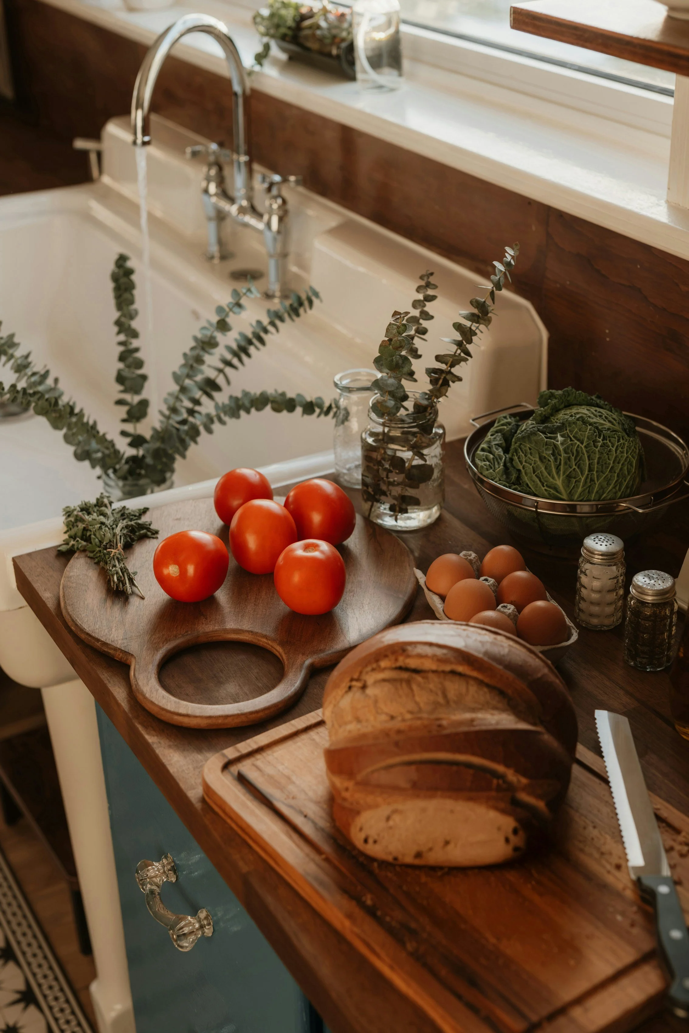 A kitchen countertop with fresh tomatoes, eggs, a loaf of bread, lettuce, and herbs near a sink, with kitchen utensils and jars nearby.