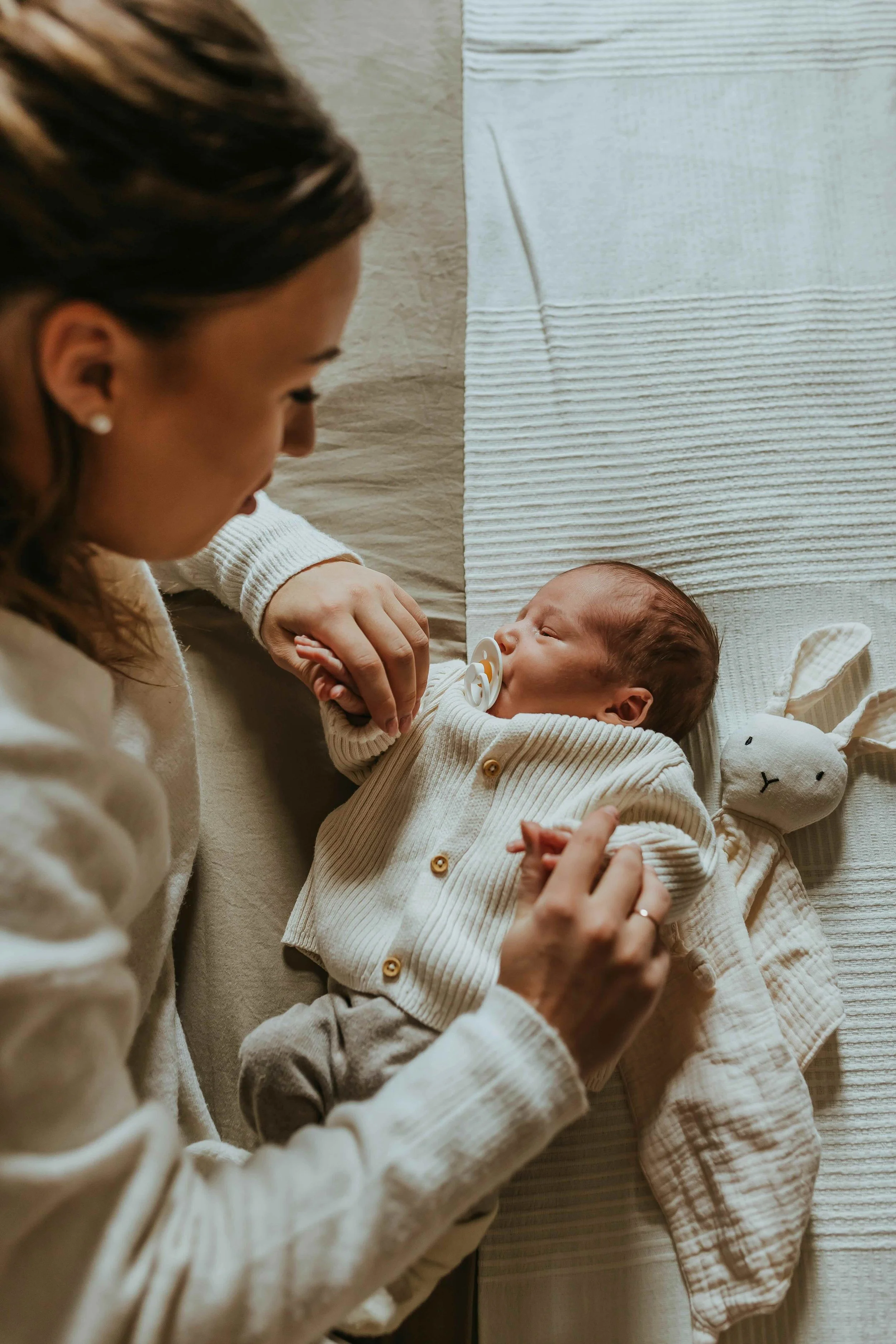 A woman gently holding a baby with a pacifier, lying on a change table with a plush bunny toy nearby.