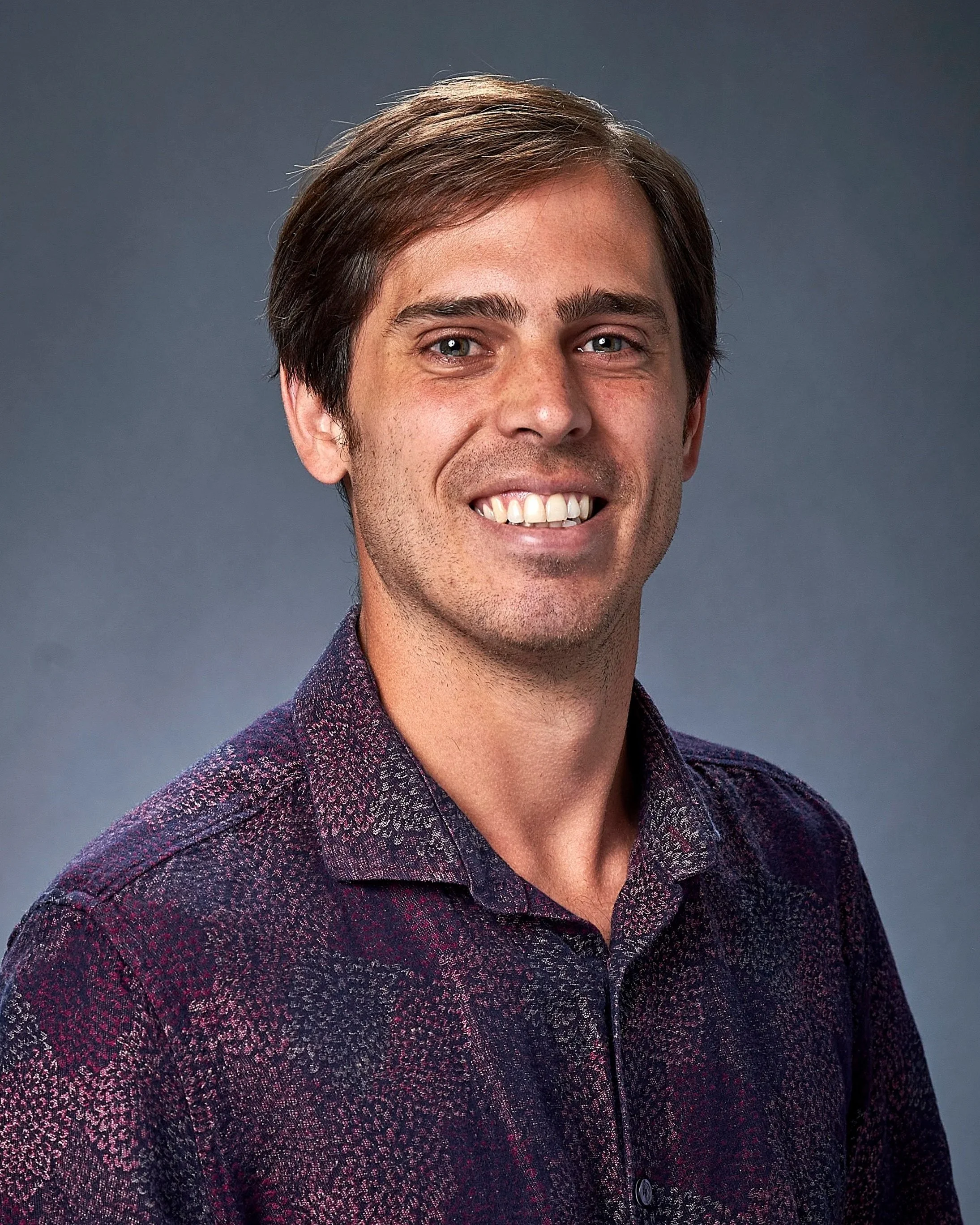 A smiling man with brown hair and blue eyes, wearing a patterned button-up shirt, against a gray background.