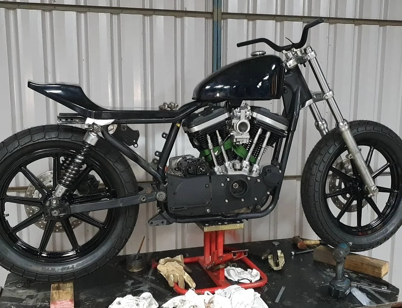 A black motorcycle is on a work stand inside a garage with metal panel walls, surrounded by tools and cloths.