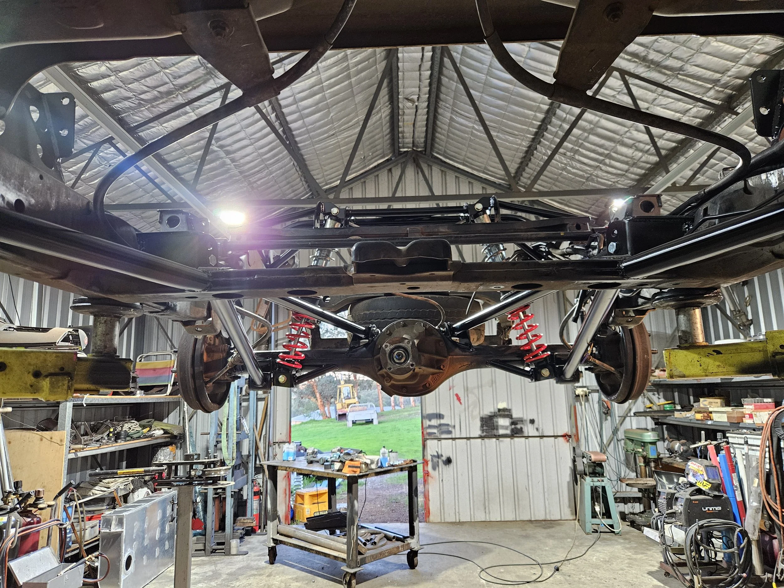 Inside an automotive workshop, a vehicle's chassis is suspended in the air with various tools and equipment visible around.