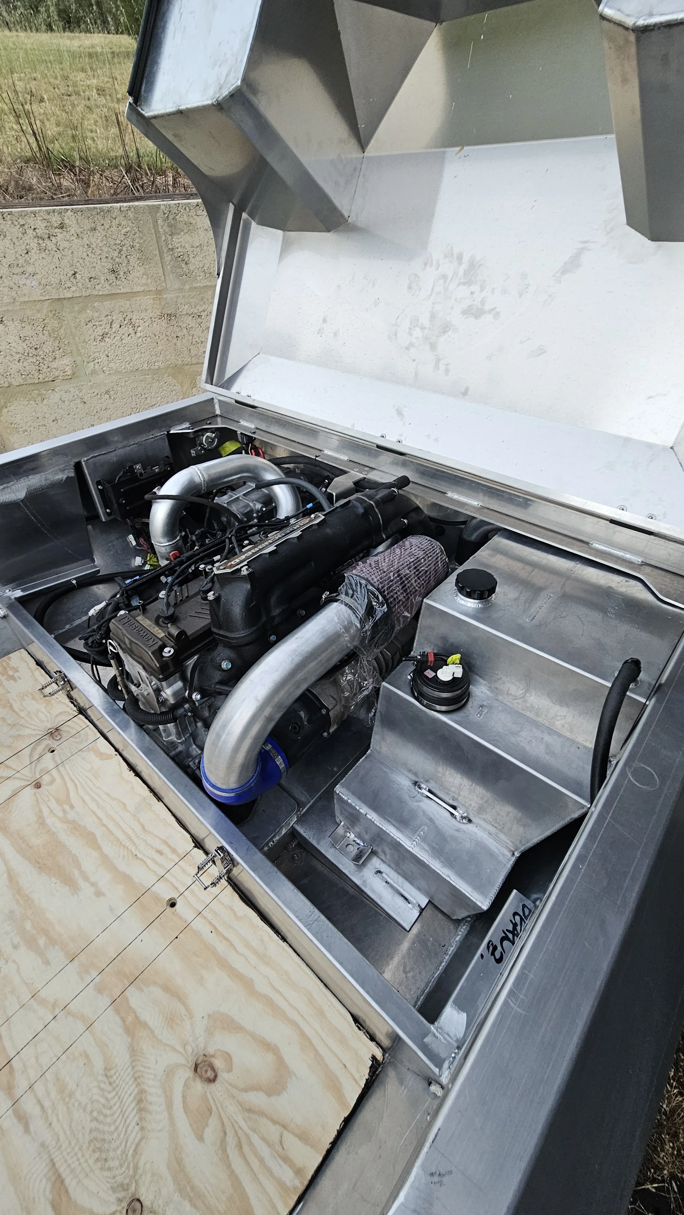 Close-up of a race car engine inside a metal compartment with an open lid, showing components and a silver intake pipe, with a wooden panel partially covering the front.