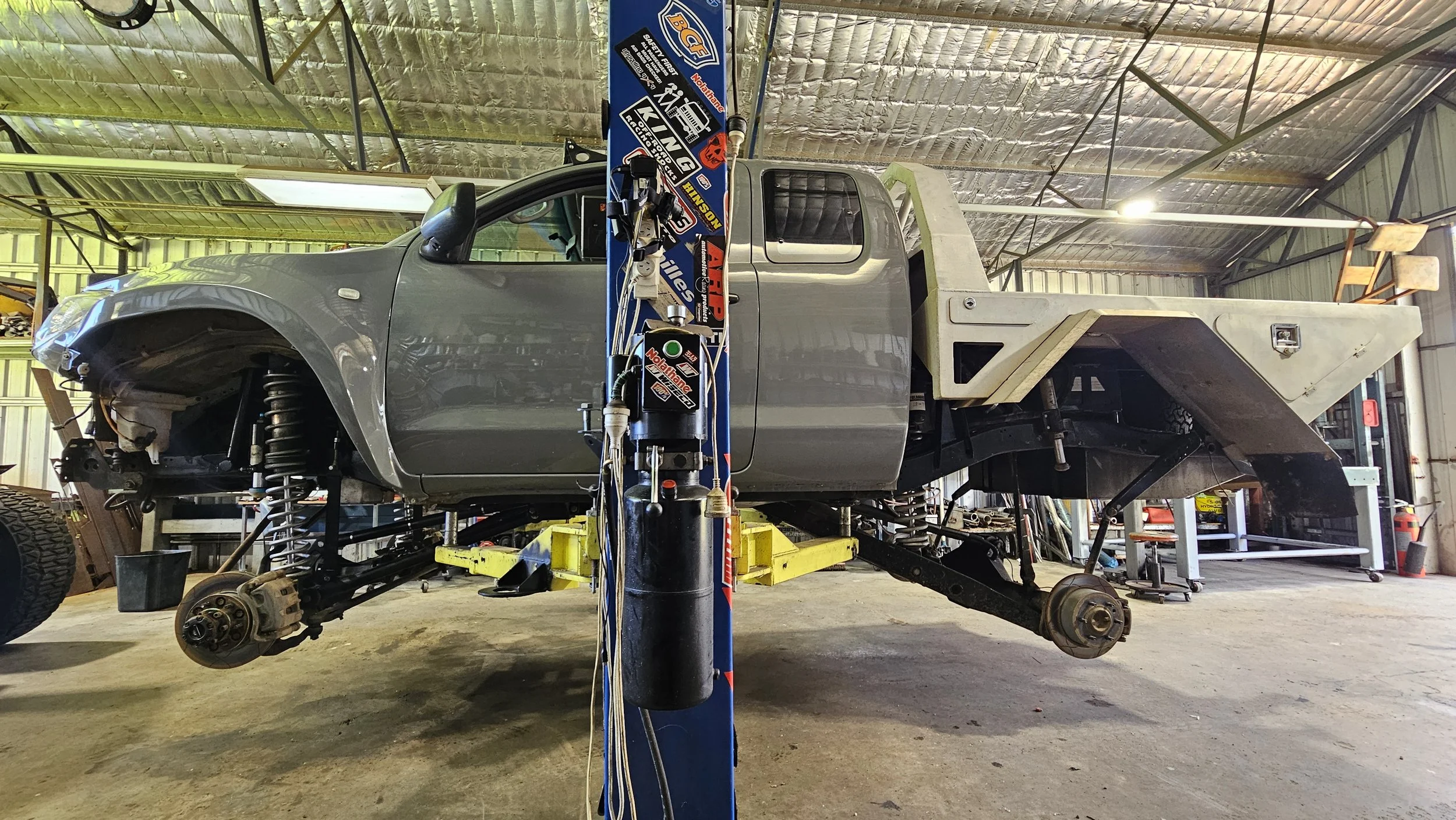 A gray pickup truck in a workshop, elevated on a hydraulic lift, with the front wheels removed and parts of the suspension visible. The truck appears to be under maintenance or repair.