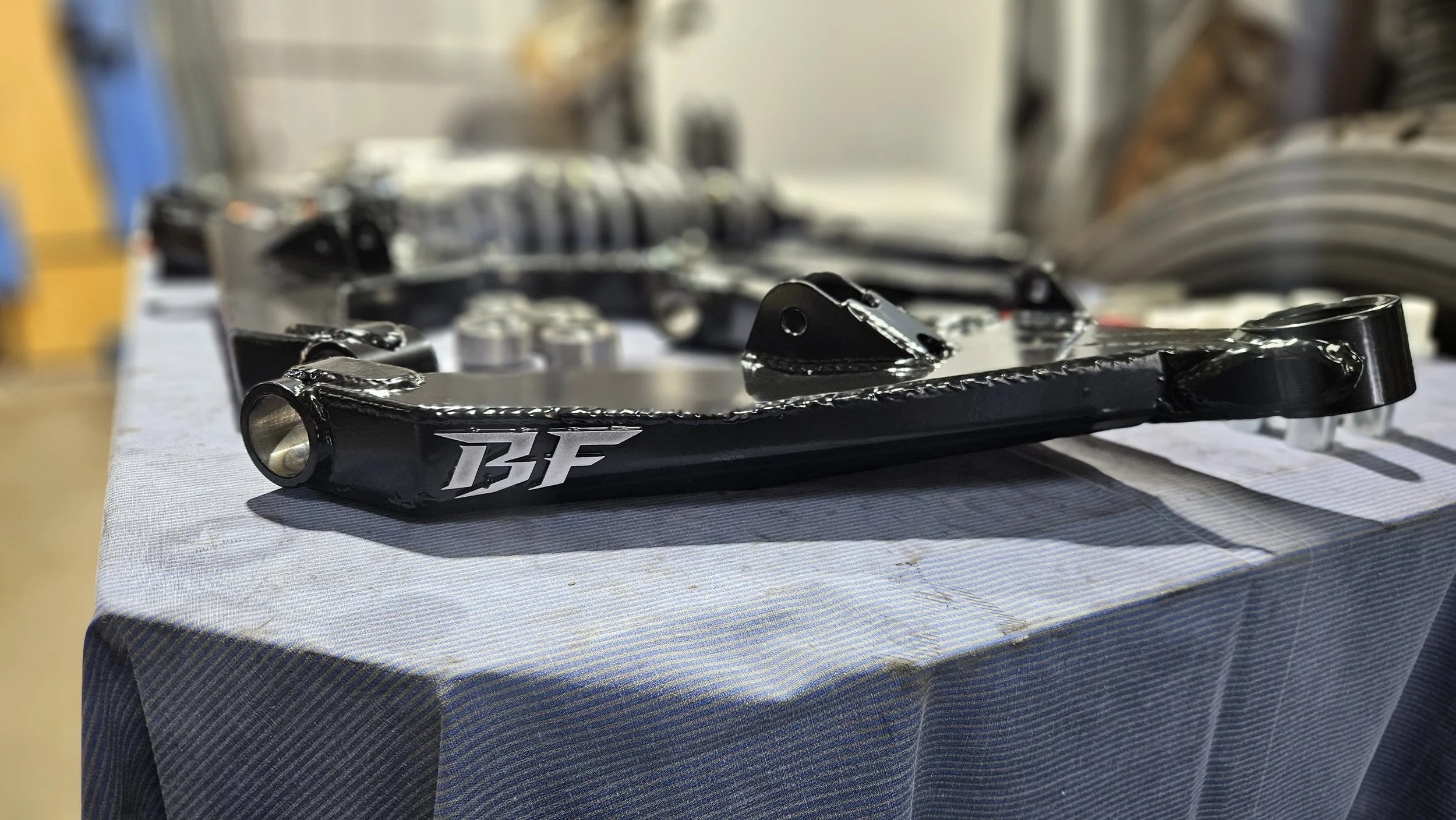 Close-up of a black automotive suspension arm with the letters 'BF' on it, resting on a table with a striped tablecloth, in a workshop or garage setting.