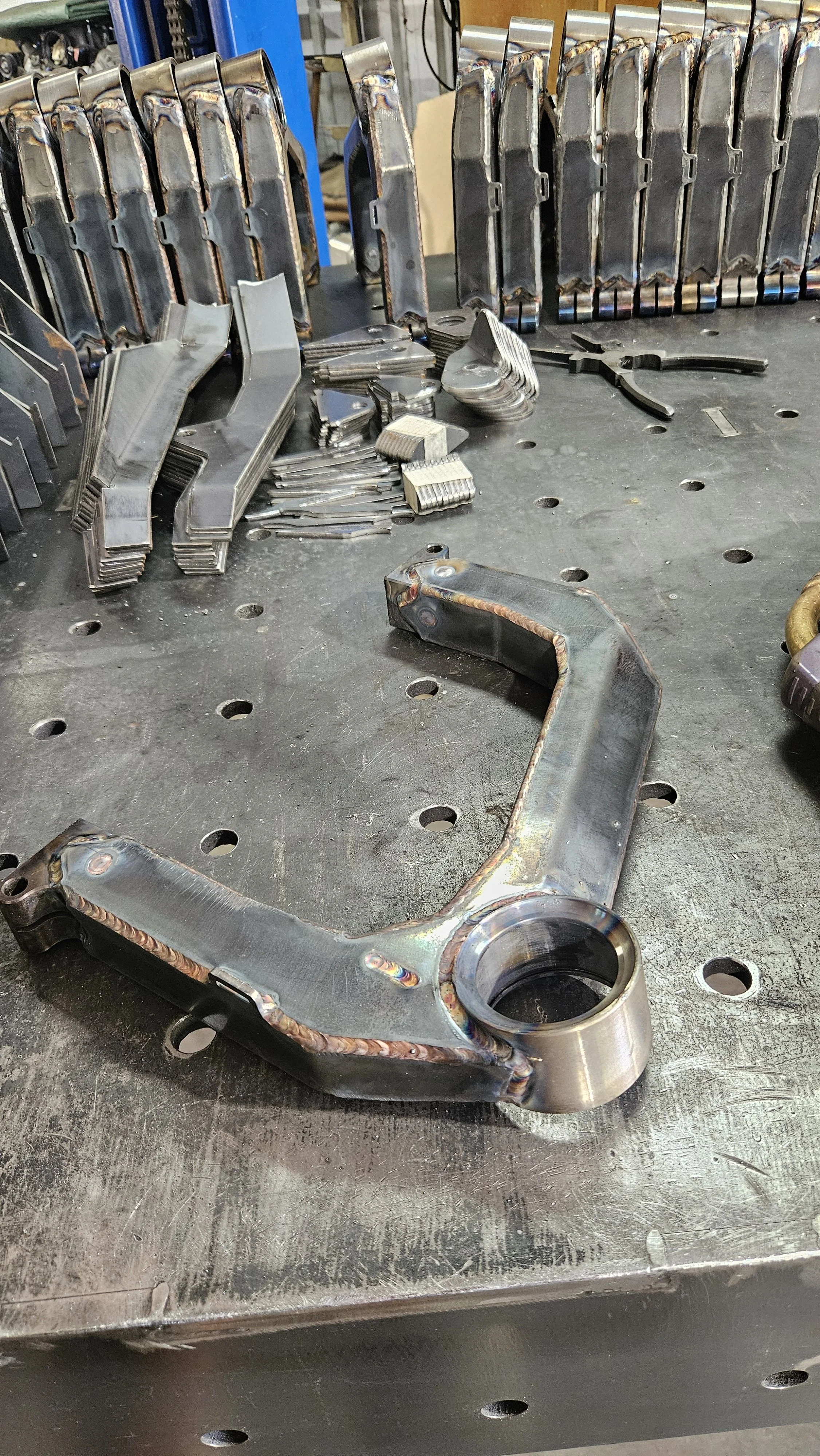 A metal automotive component with welded seams, placed on a workbench with other metal parts and tools in the background.