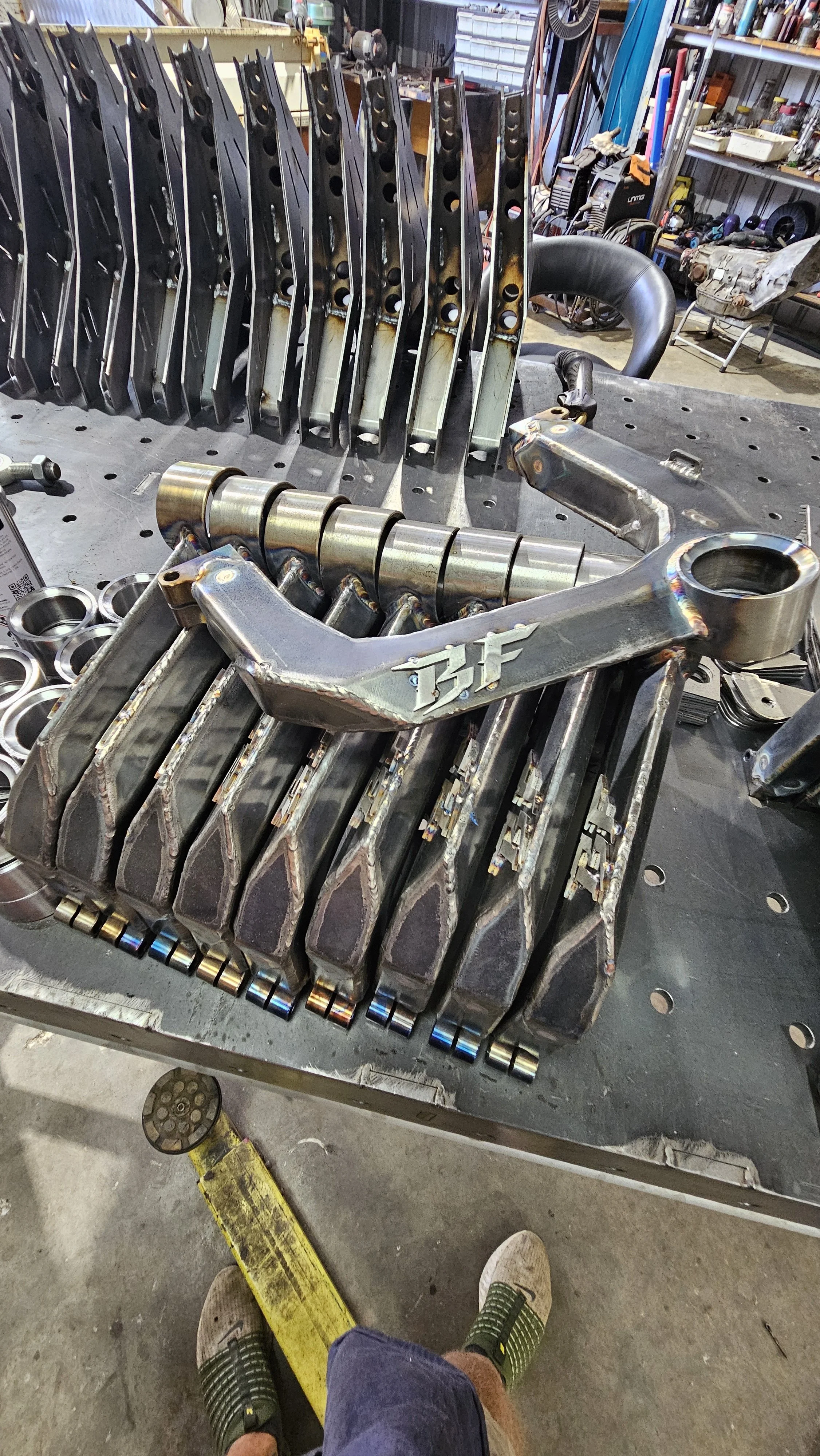 A workbench with various automotive parts, including metal brackets, a camshaft, and other engine components, in a workshop setting.