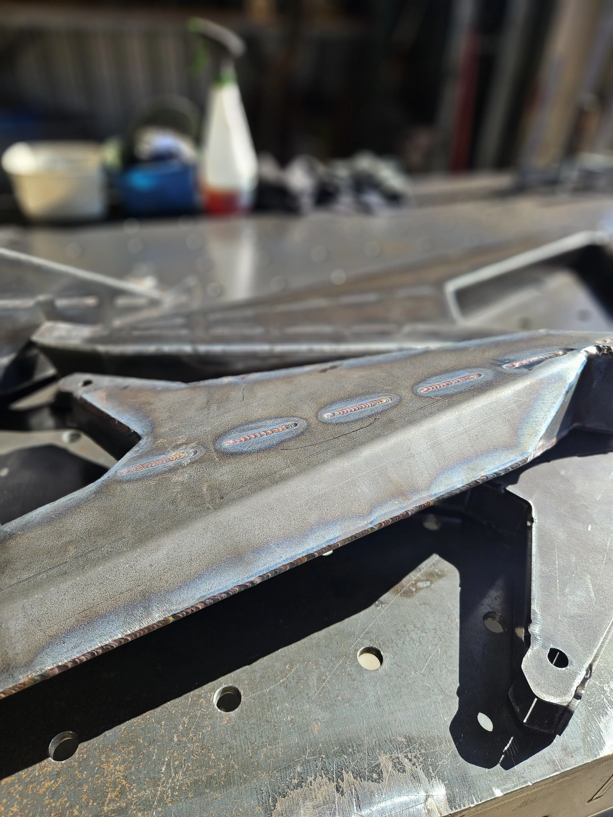 Close-up of a metal piece with rainbow-colored welds on a workbench, with blurred tools and supplies in the background.