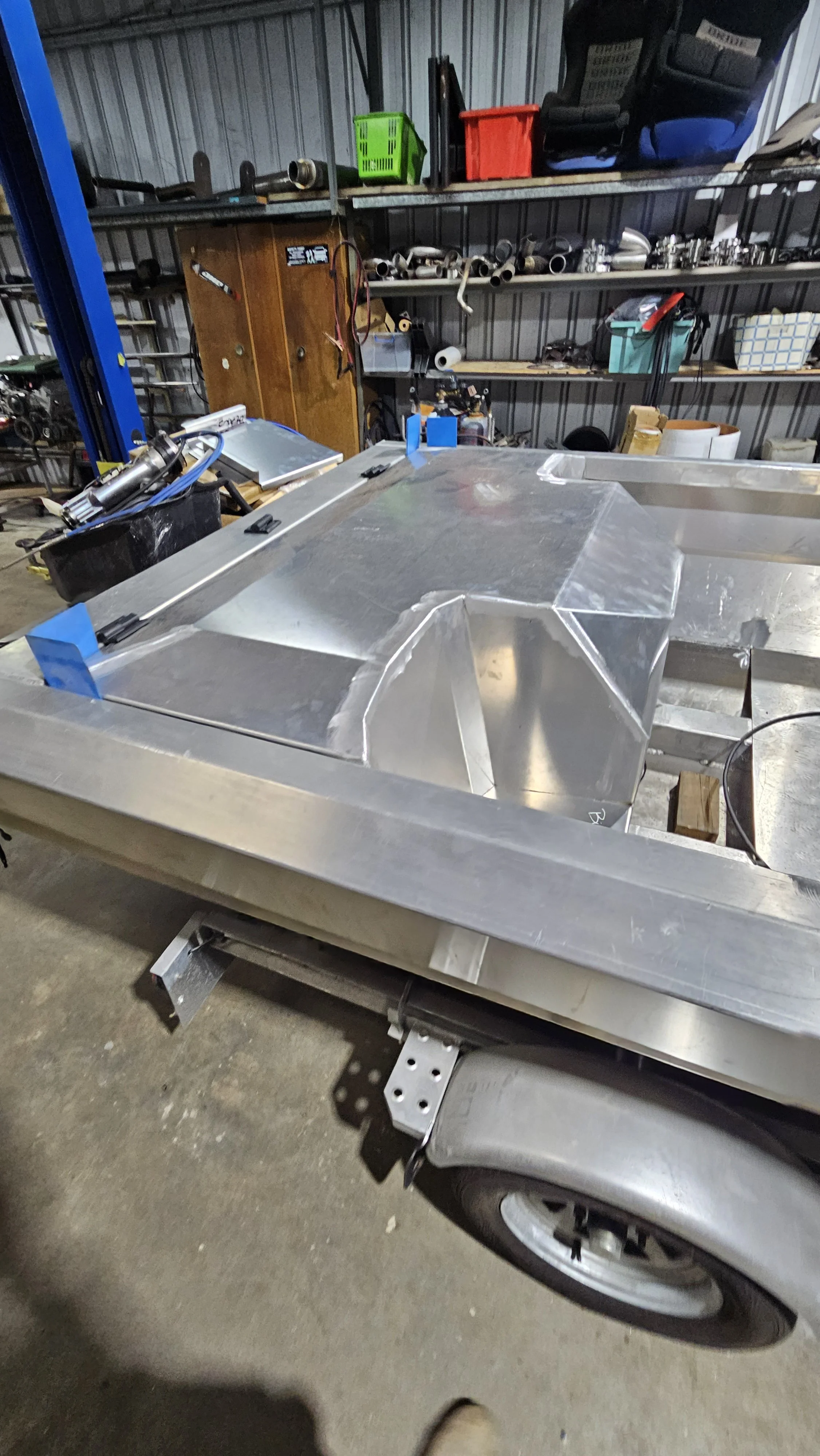 A metal work table with an aluminum frame and components, in a workshop setting with shelves holding tools, parts, and containers in the background.