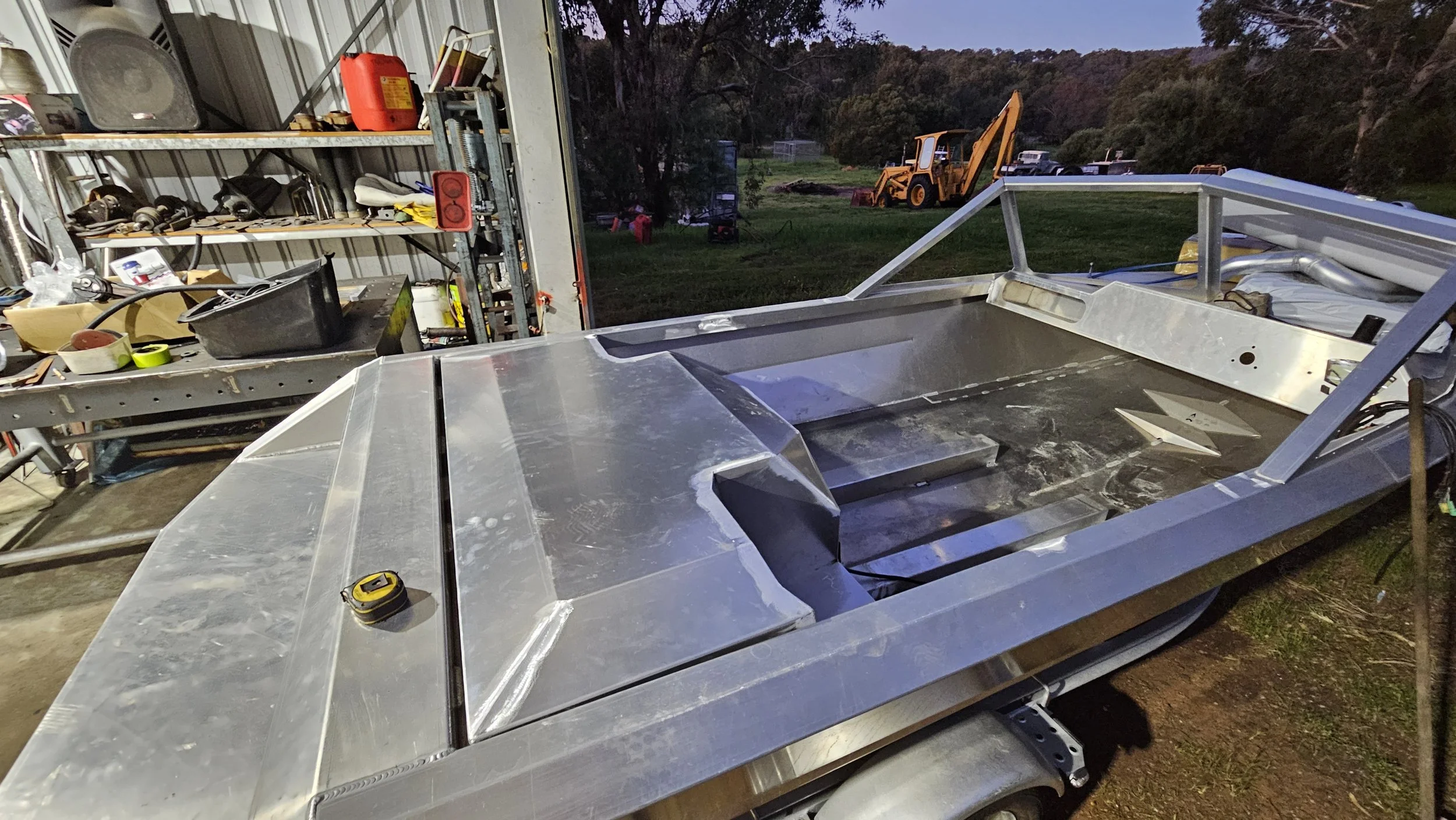An unfinished aluminum boat on a trailer inside a workshop, with a workbench and shelves filled with tools and supplies in the background, and outdoor equipment visible through an open door at dusk.