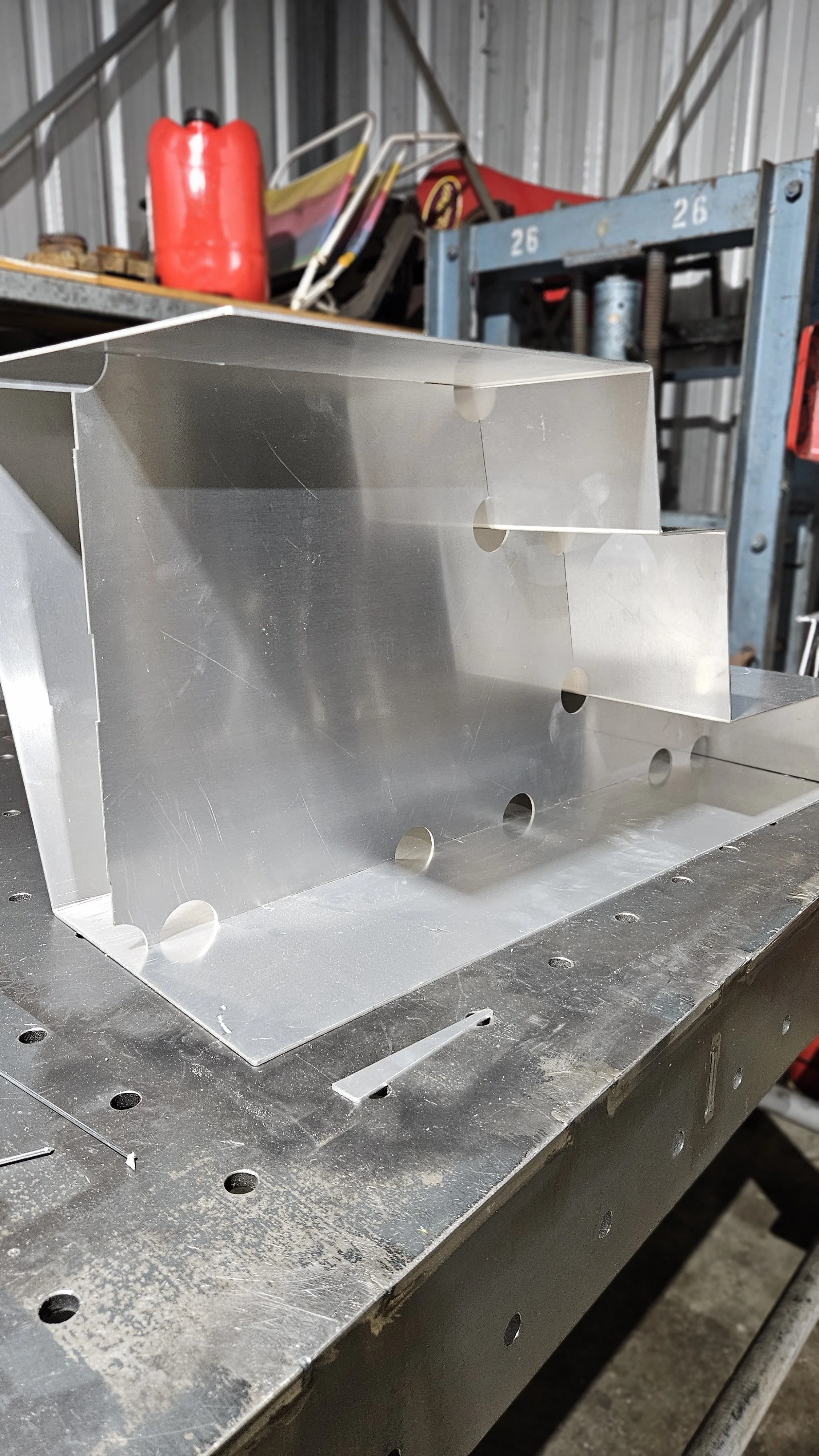 A metal fabrication work in progress on a workbench in an industrial workshop, with a metal enclosure, a red container, and workshop tools in the background.