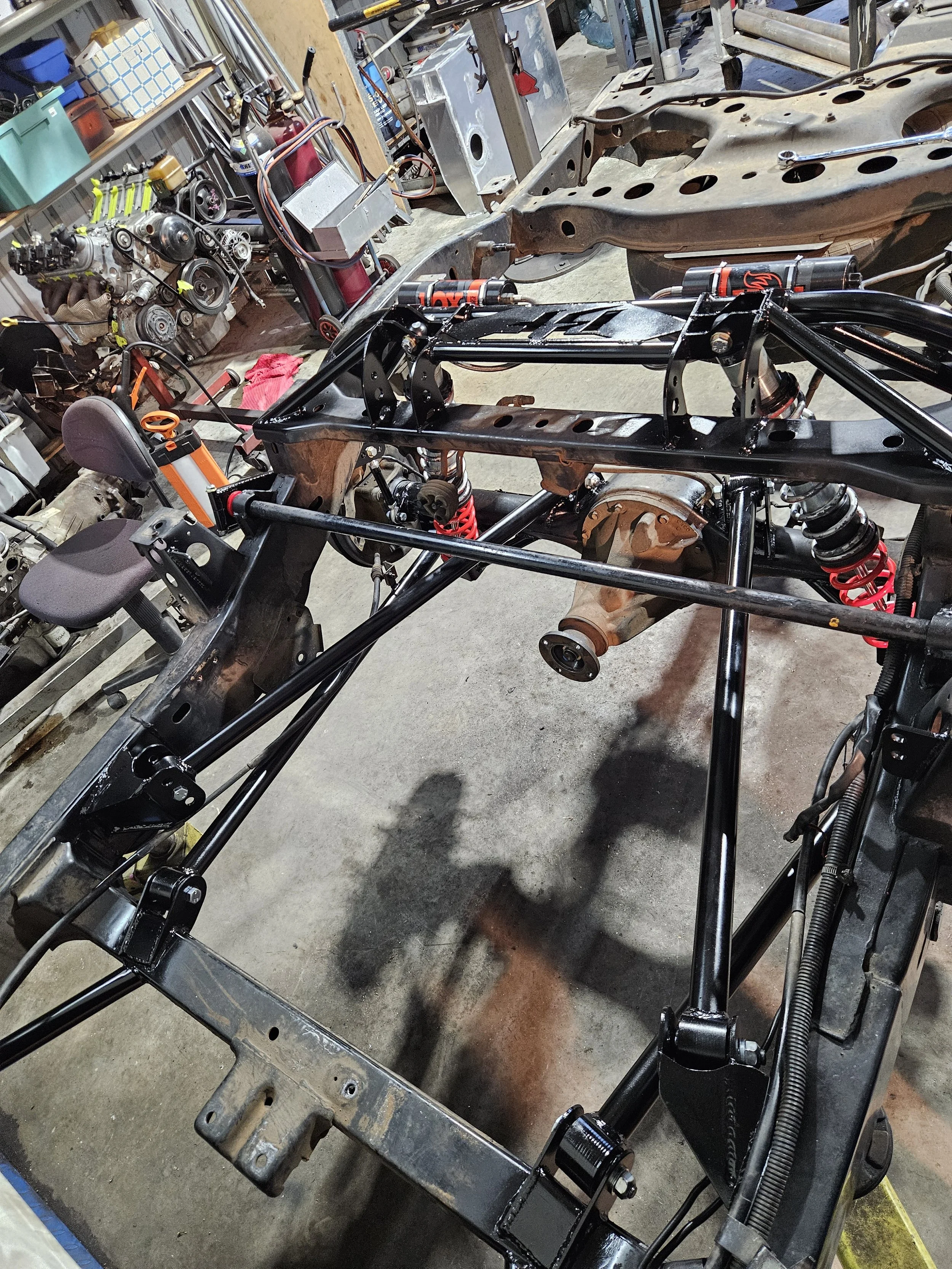 The image shows a partially assembled car chassis in a workshop, with some suspension components, shocks, and a steering seat visible. Various tools and auto parts are scattered around in the background.