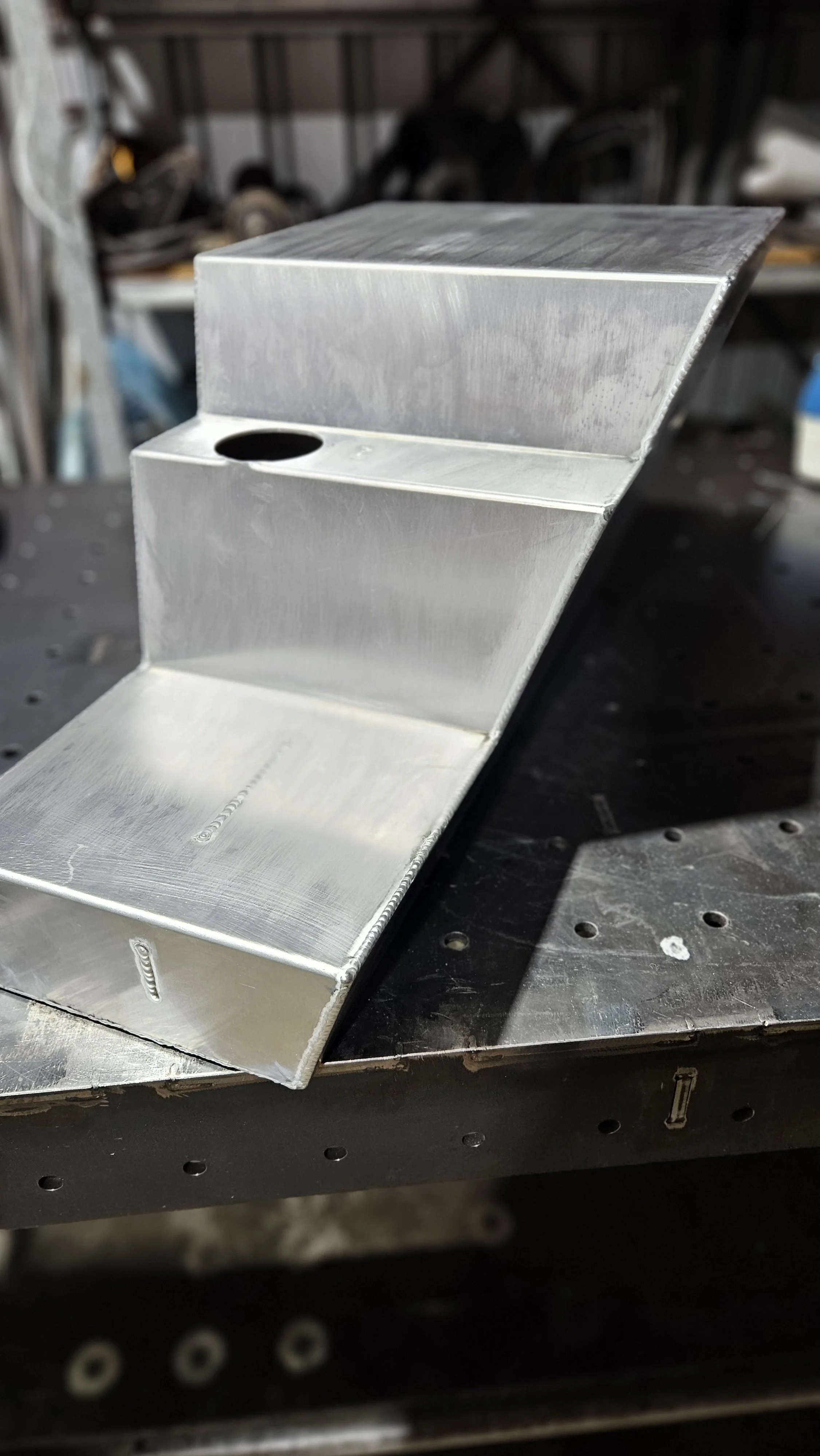 A metal bracket with two stepped sections and a circular hole, placed on a black workbench in a workshop.