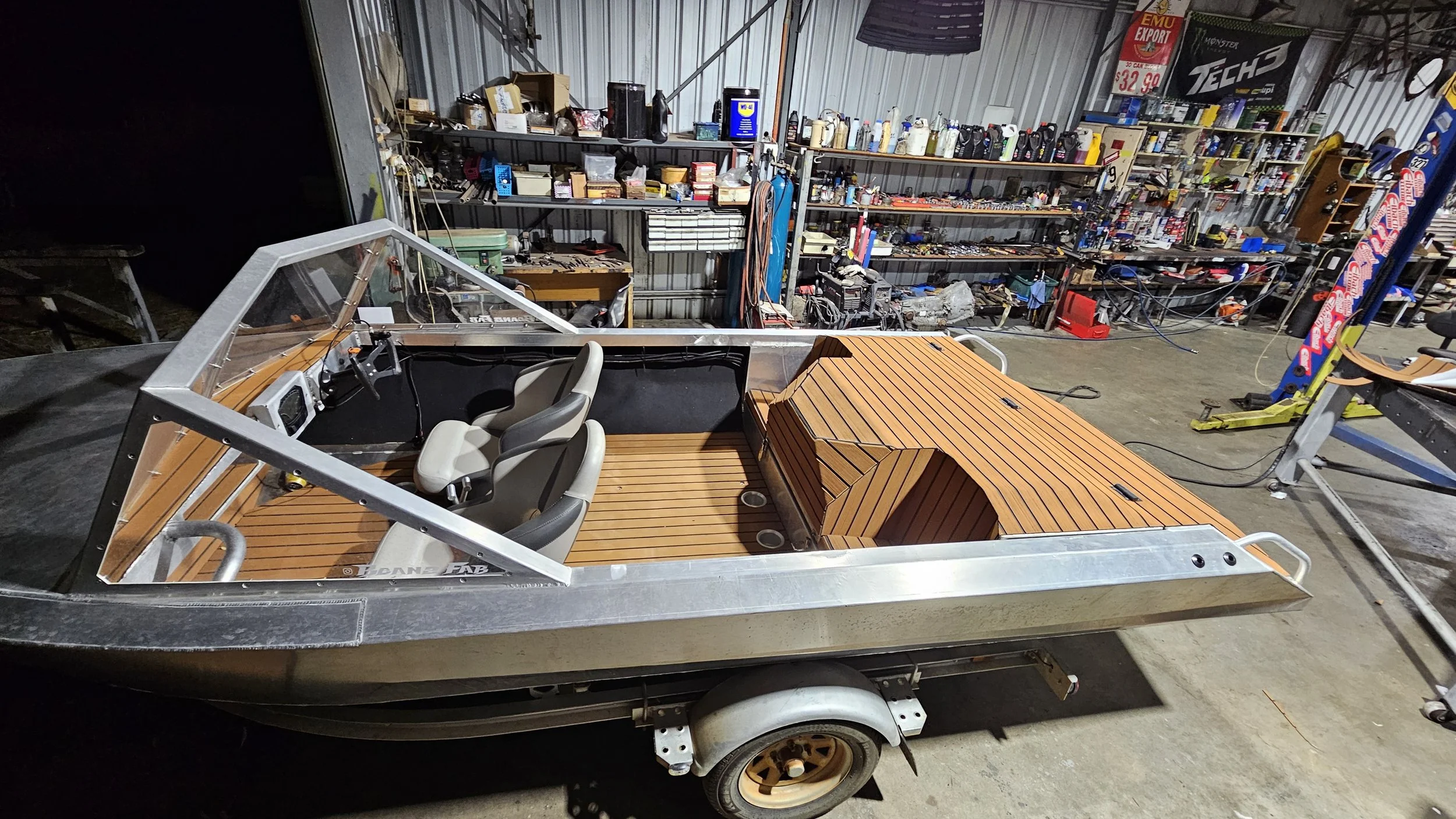 A partially assembled boat with a wooden deck and three beige seats inside a workshop or garage with shelves filled with tools and equipment in the background.