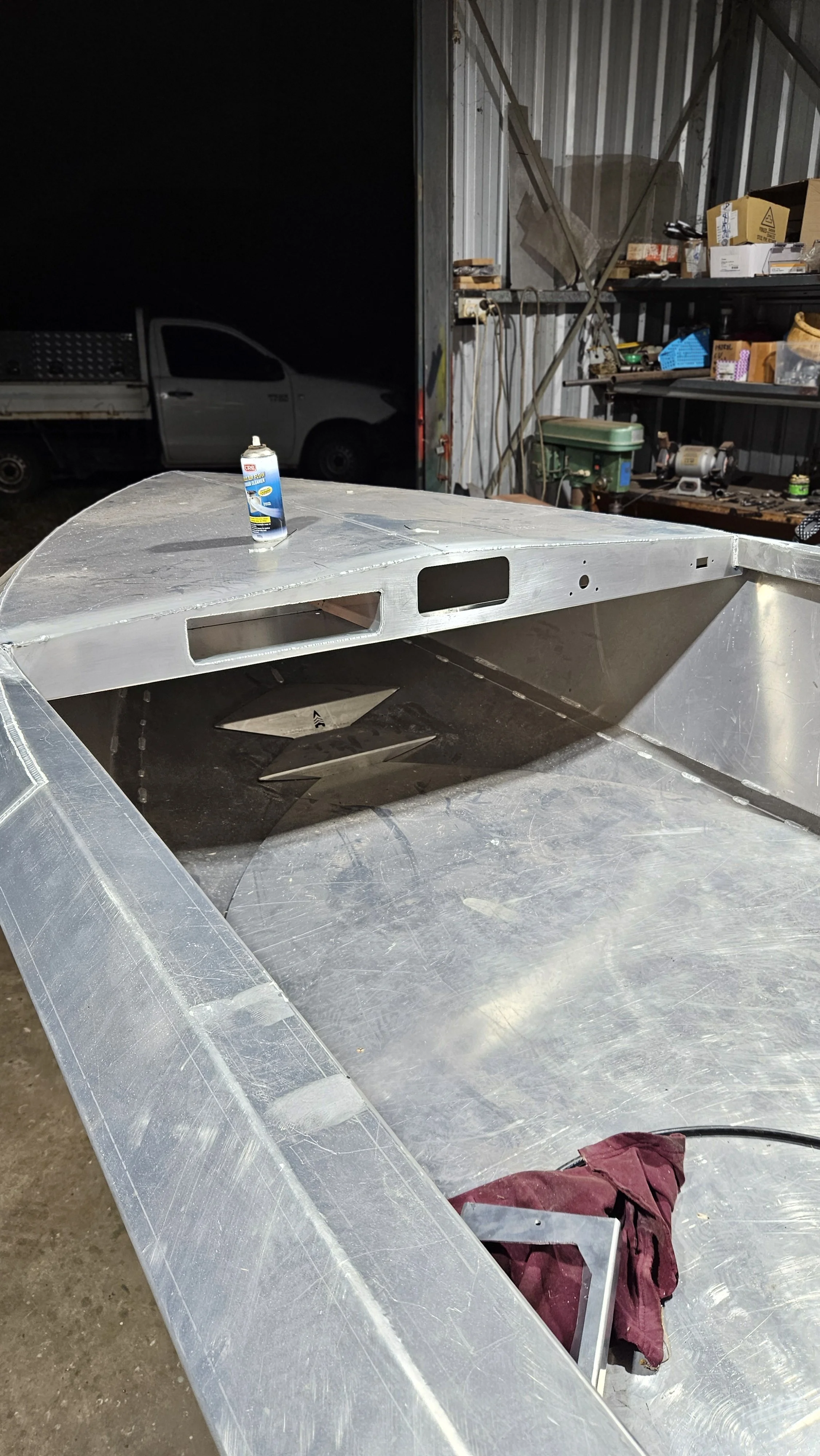 Close-up of a metal boat or aircraft fuselage under construction in a workshop, with a can of spray adhesive on top, workshop tools and shelves in the background.