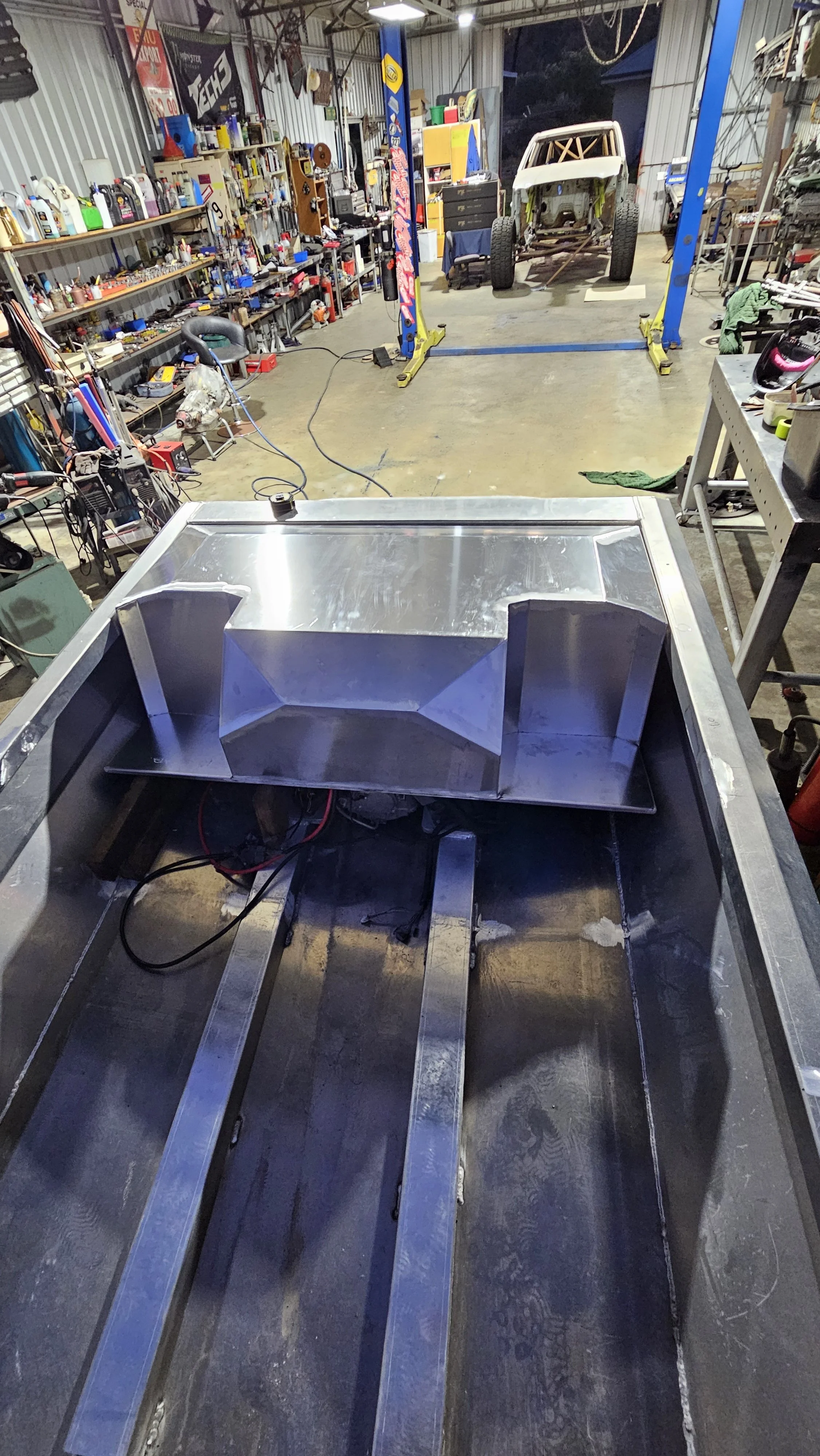 View of a metal fabrication workshop with a partially assembled metal structure in the foreground, various tools, supplies, and a vehicle frame on a lift in the background.
