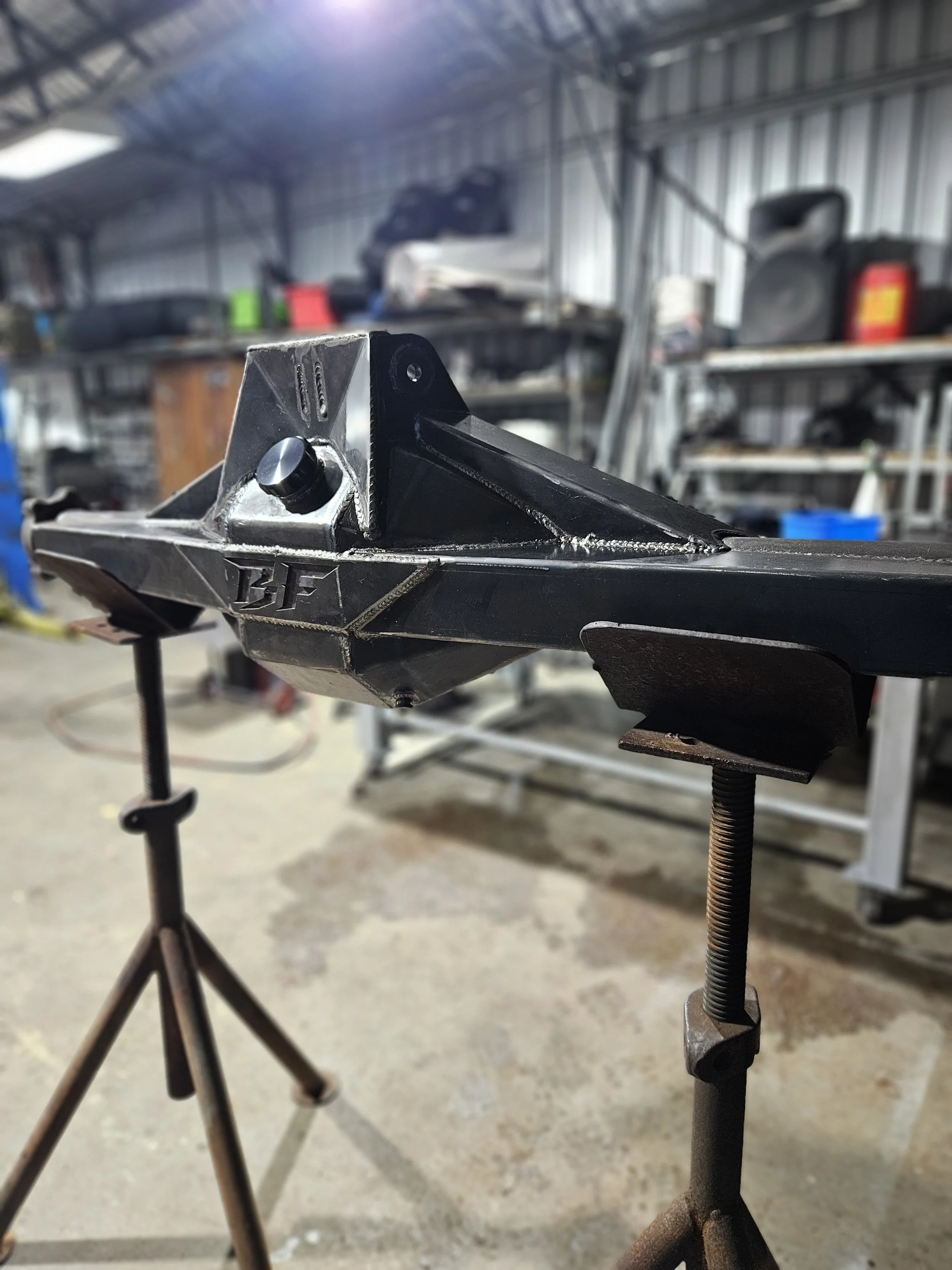 Close-up of a black metal automotive or mechanical part, clamped in place on a workbench in a workshop, with tools and shelves in the background.