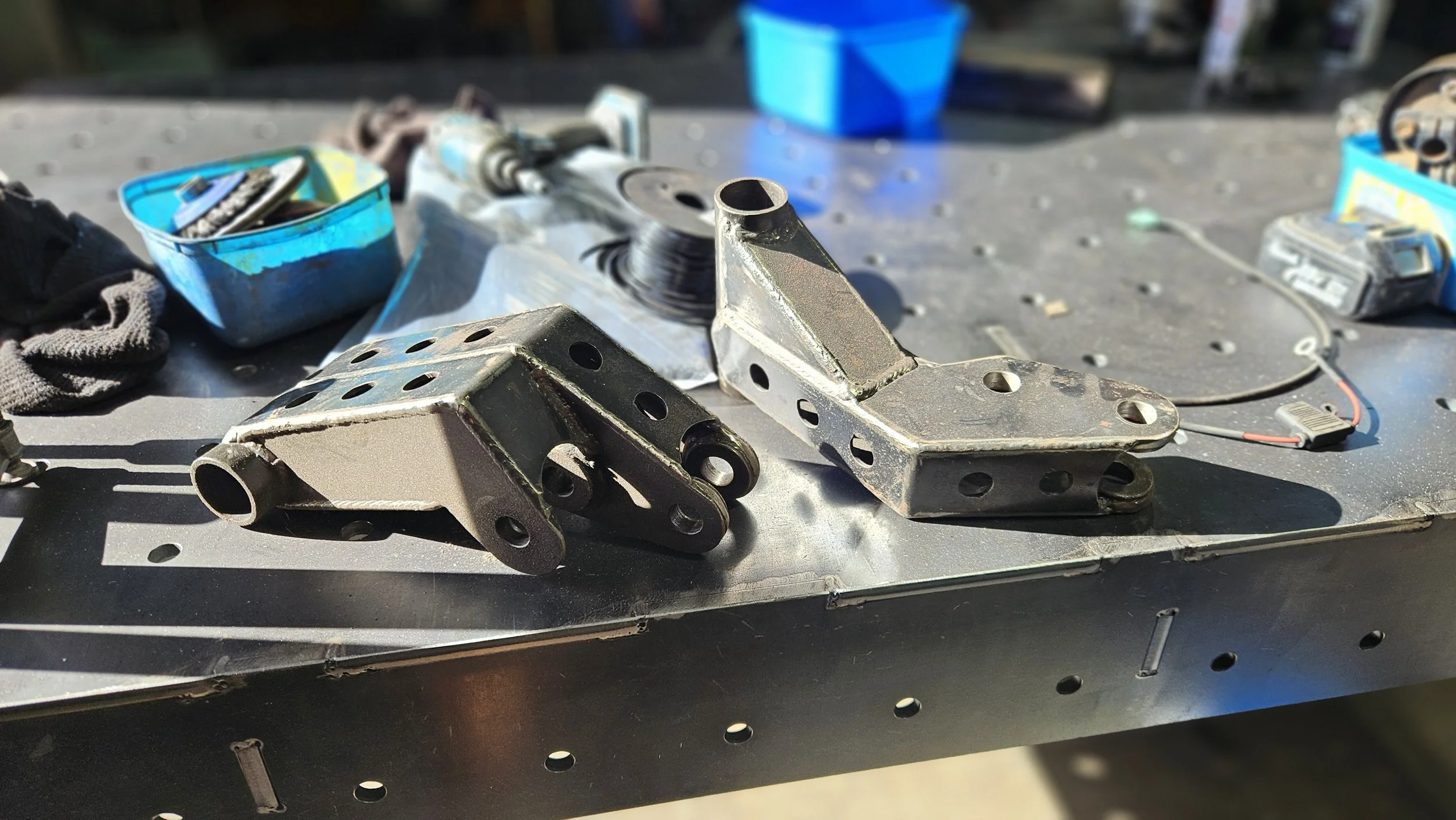 Metal brackets and mechanical parts on a workbench with plastic containers and tools in the background.