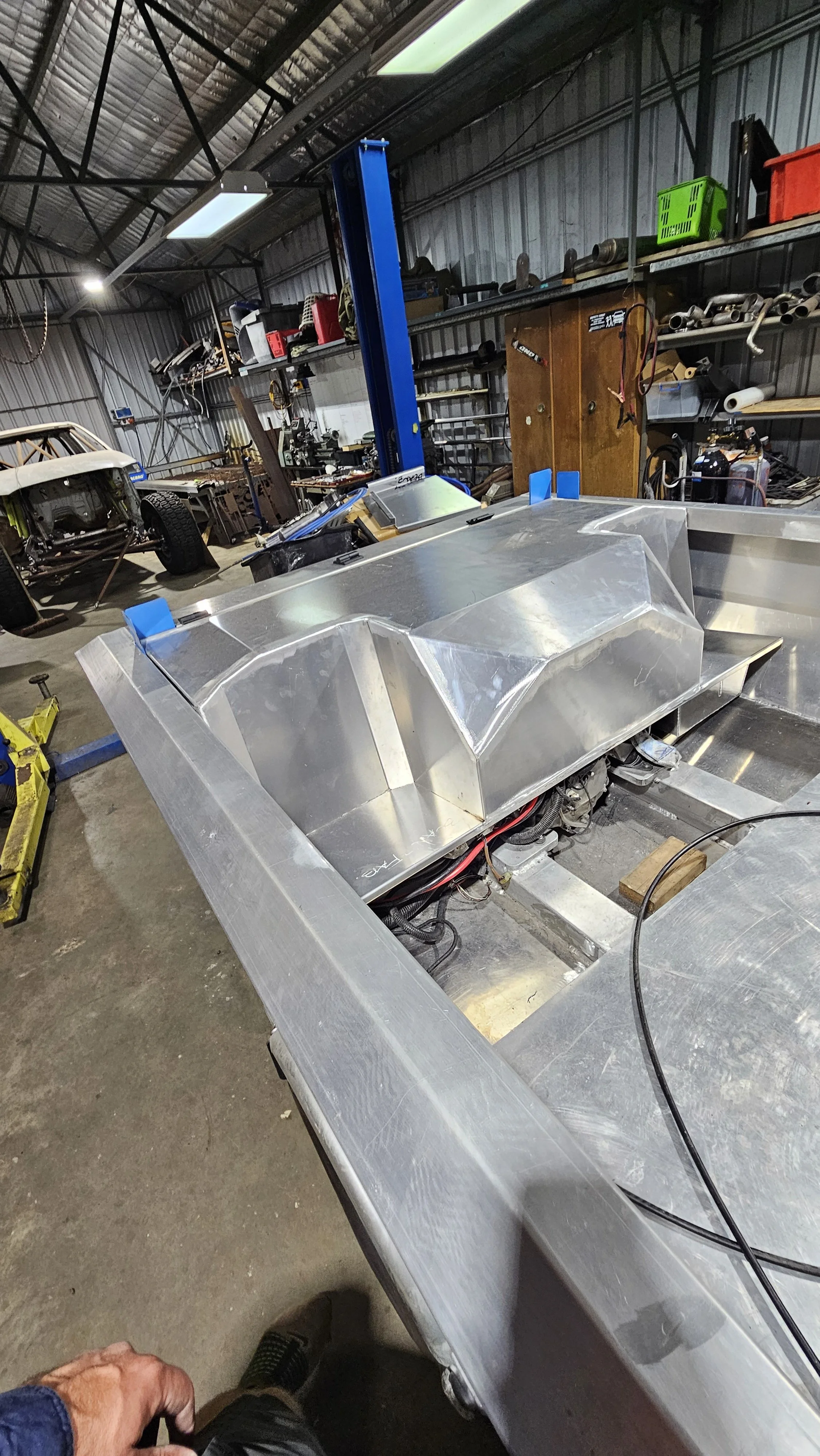 Metal frame of a race car or go-kart in a workshop, with car parts, tools, shelves, and another vehicle in the background.