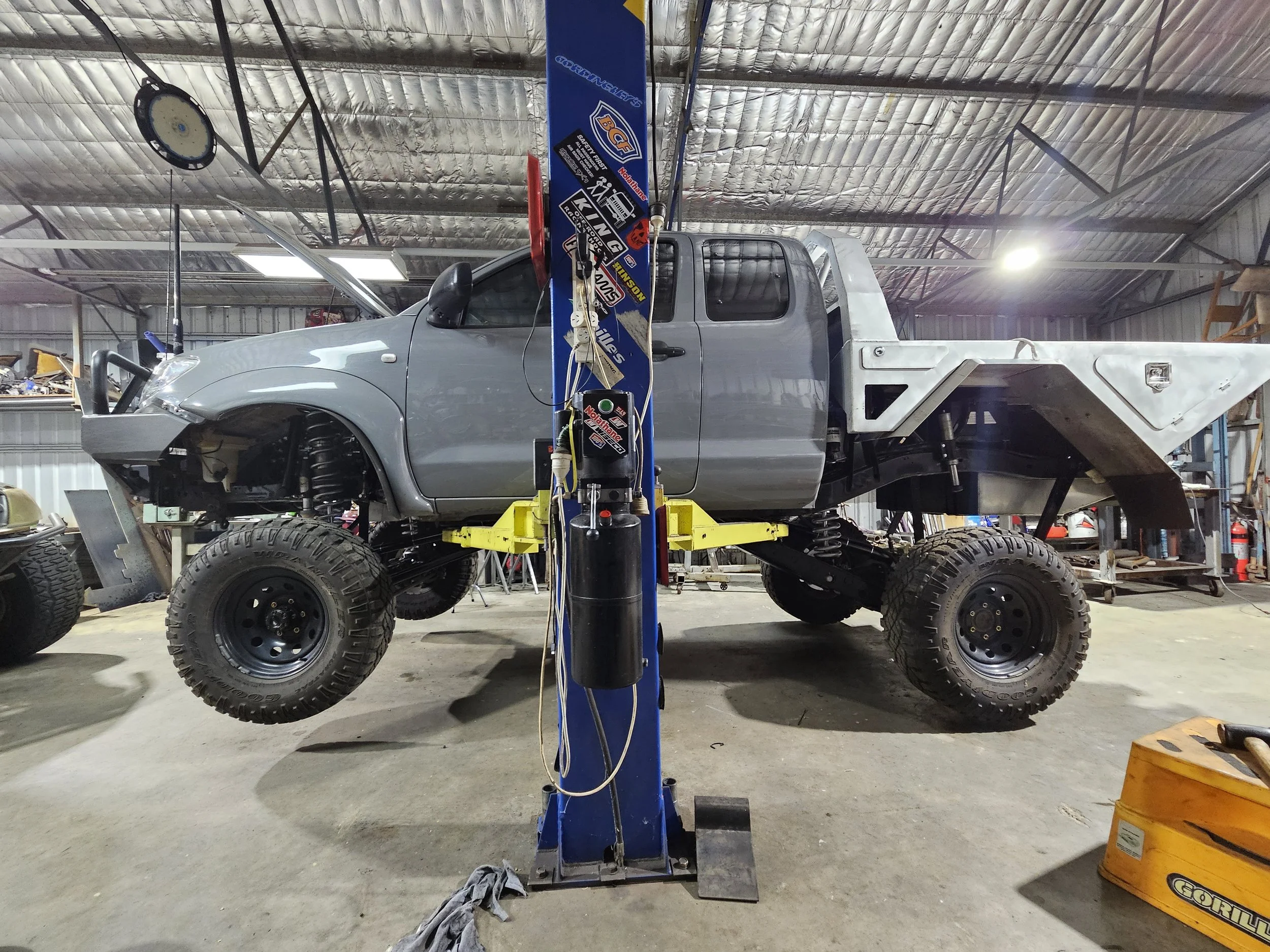 A gray pickup truck in a workshop, elevated on a lift with the front end and hood removed, exposing the suspension and engine components.