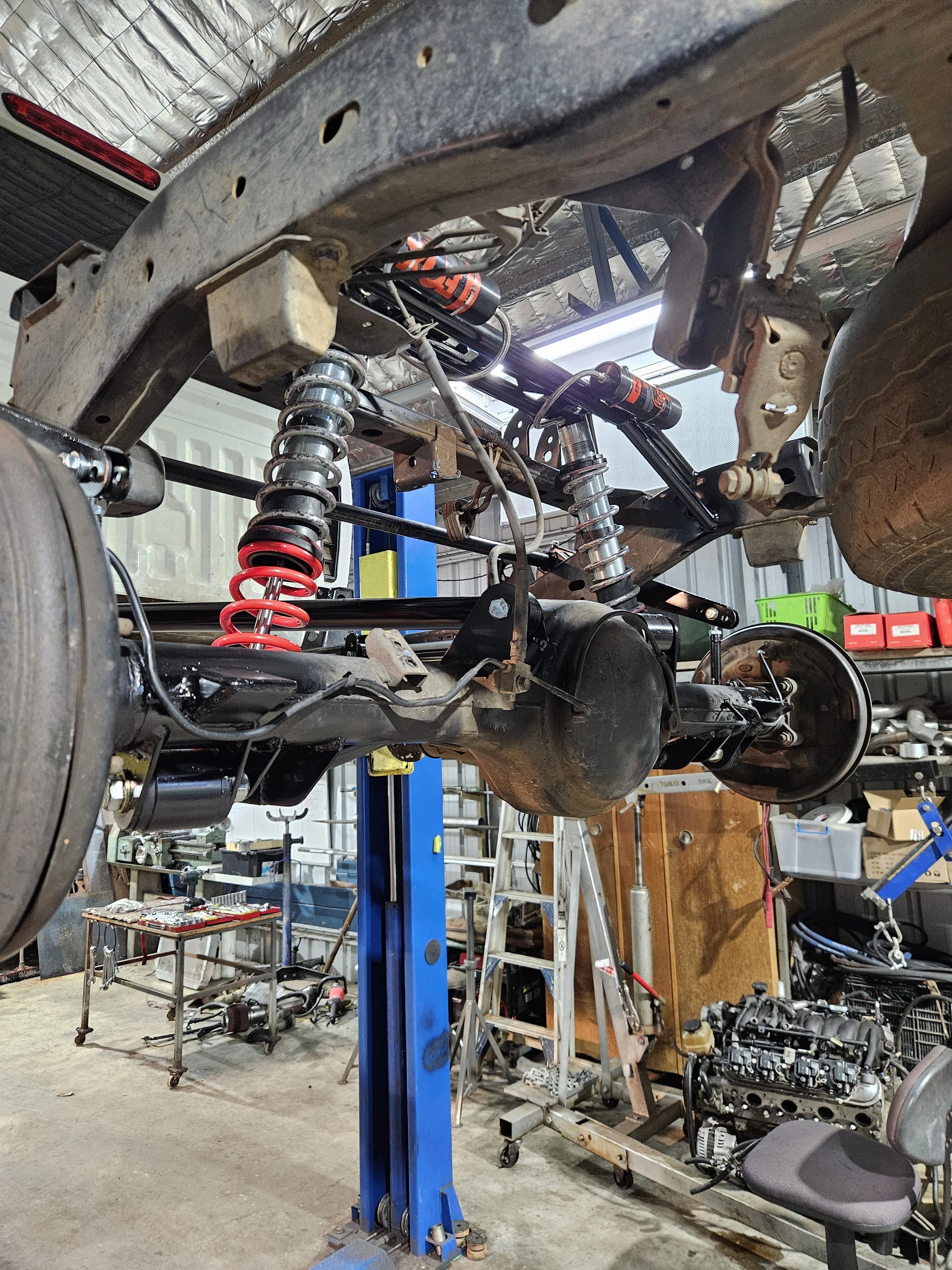 Auto repair shop with vehicle frame on a lift, showing suspension system and rear axle, surrounded by tools, a workbench, and an engine in the background.