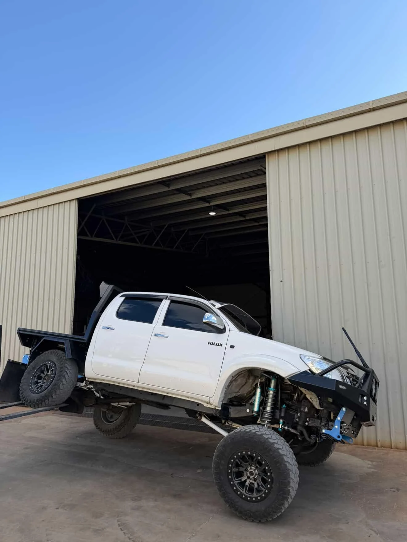 A white Toyota Hilux pickup truck with off-road modifications, including large tires, lifted suspension, and a front bumper, is parked partially outside a large metal shed with an open door.
