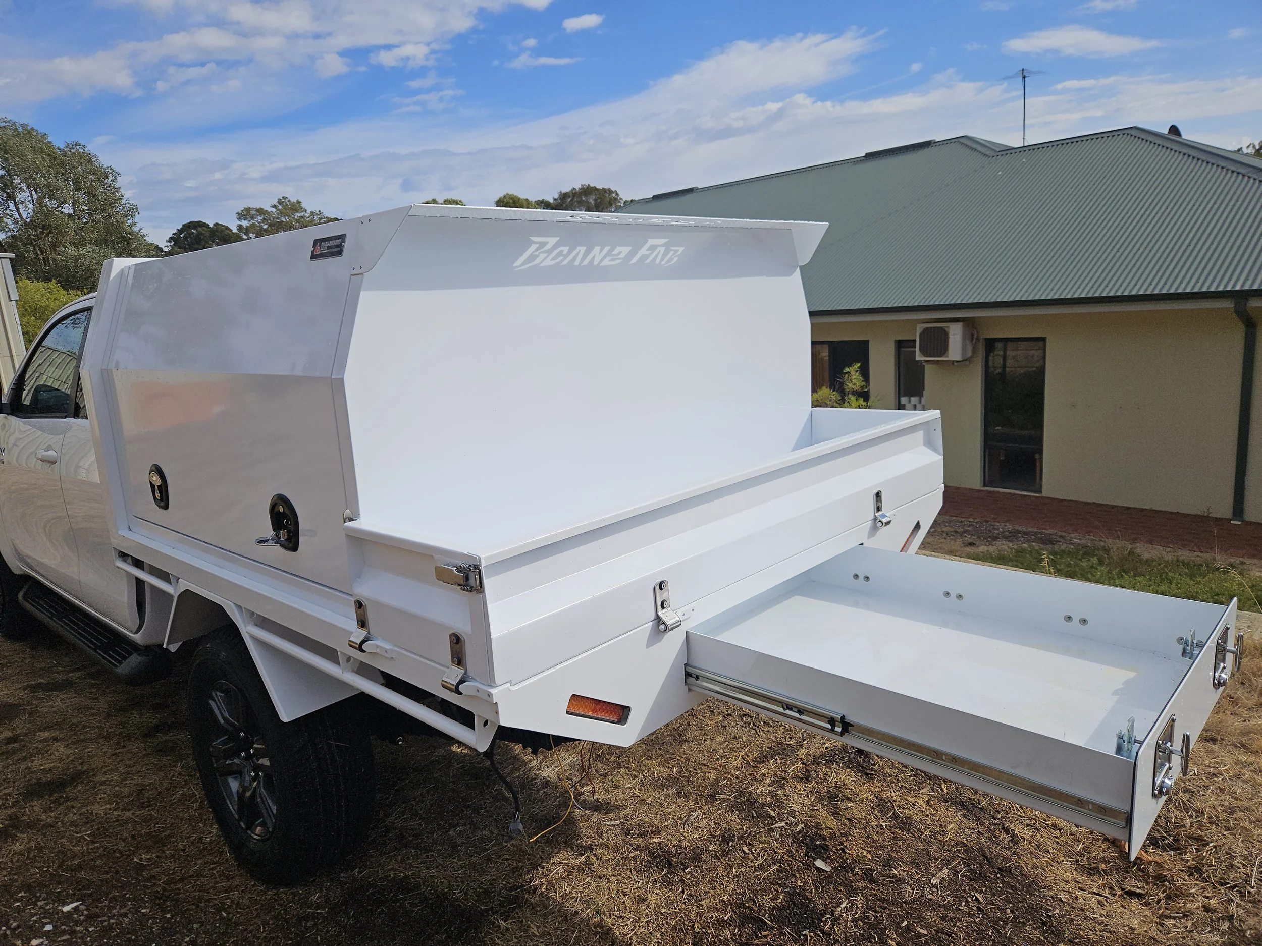 A white pickup truck with a large white utility canopy and an extension tray at the back, parked on a dirt driveway in front of a building with a green roof and an air conditioning unit.