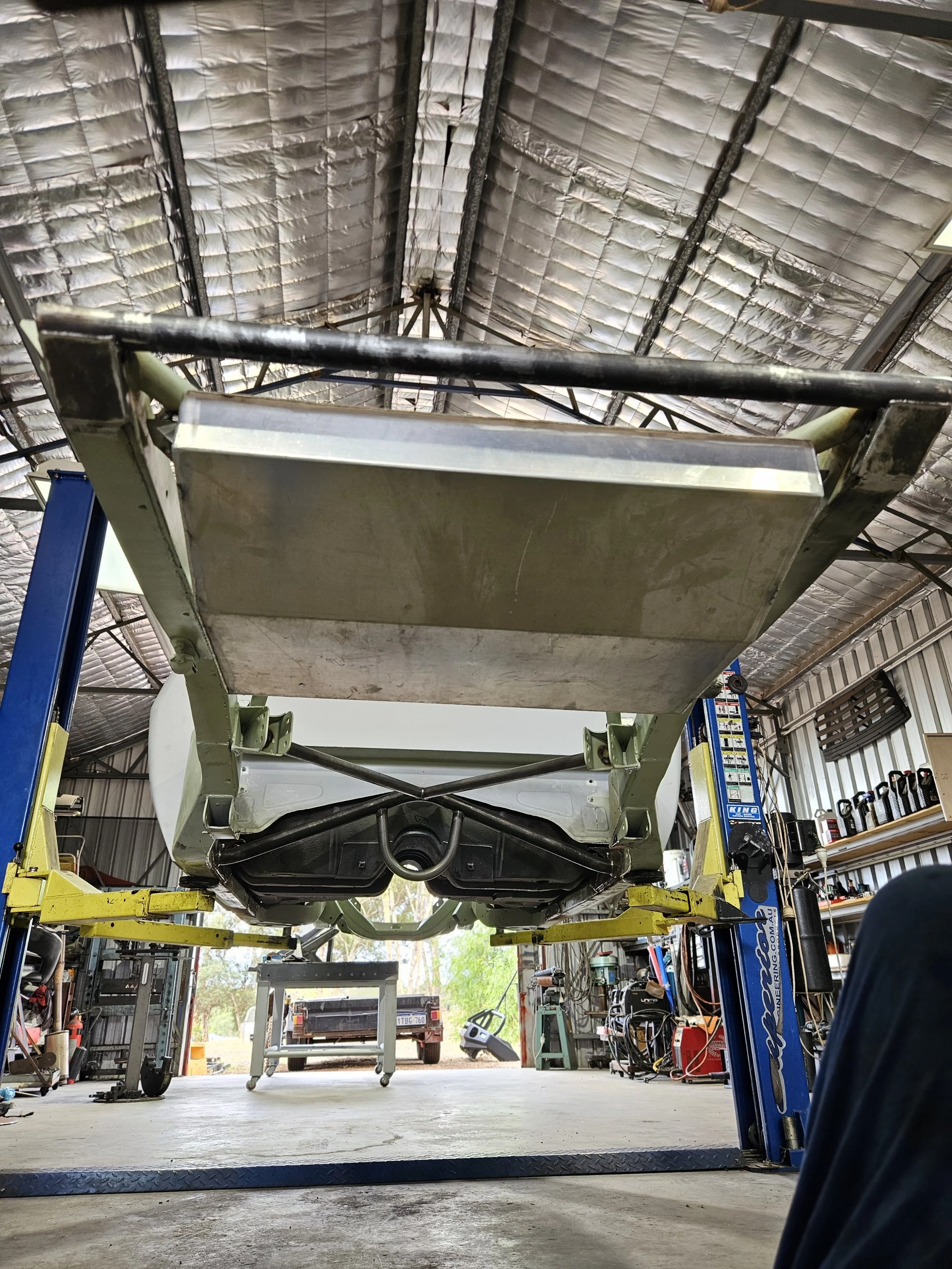 Underneath view of a car raised on a hydraulic lift inside a garage or workshop with tools and equipment on shelves.
