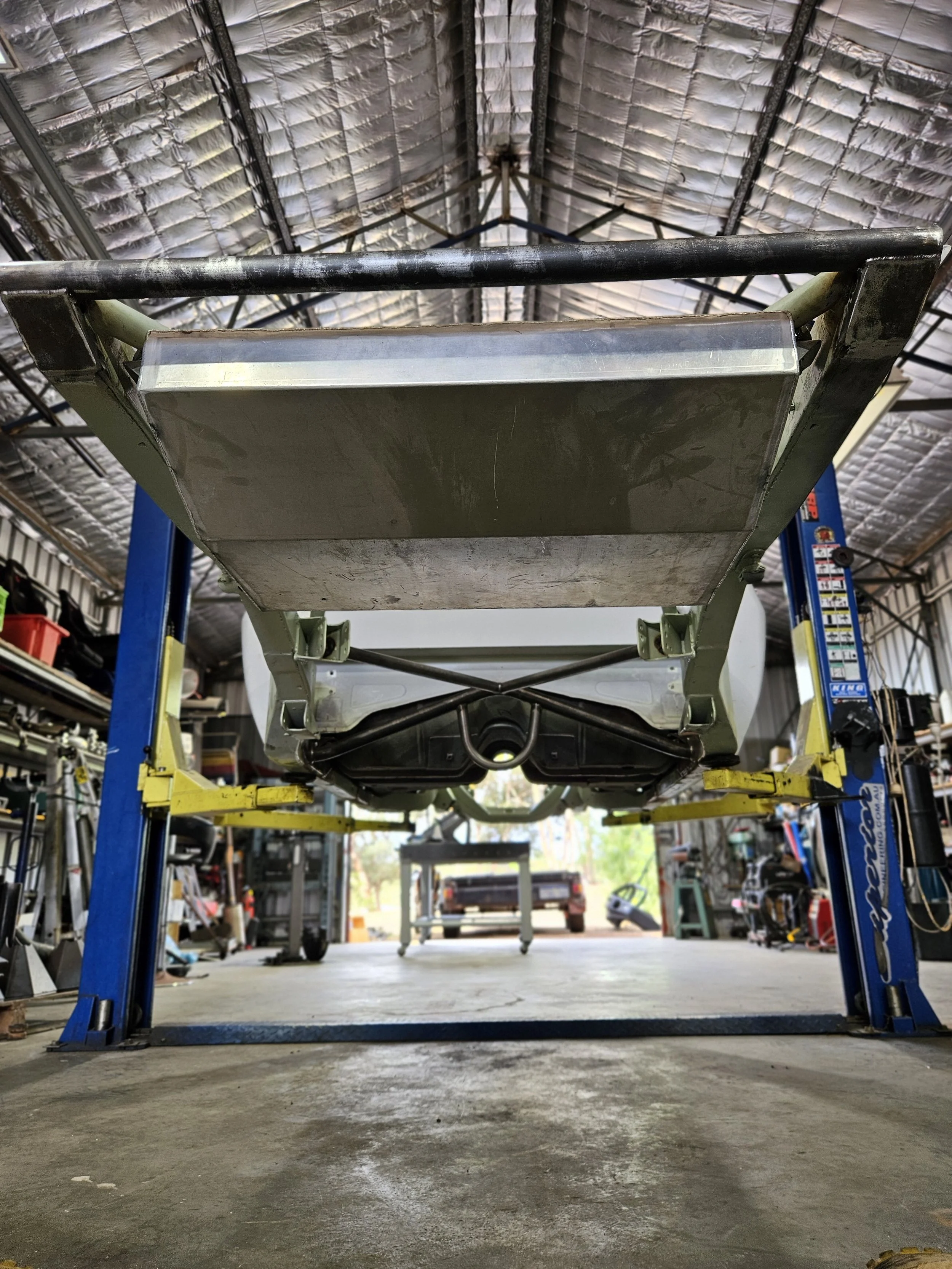 View from below of a vehicle on a hydraulic lift inside a garage, showing the underside of the vehicle, the garage ceiling, and some tools and equipment.