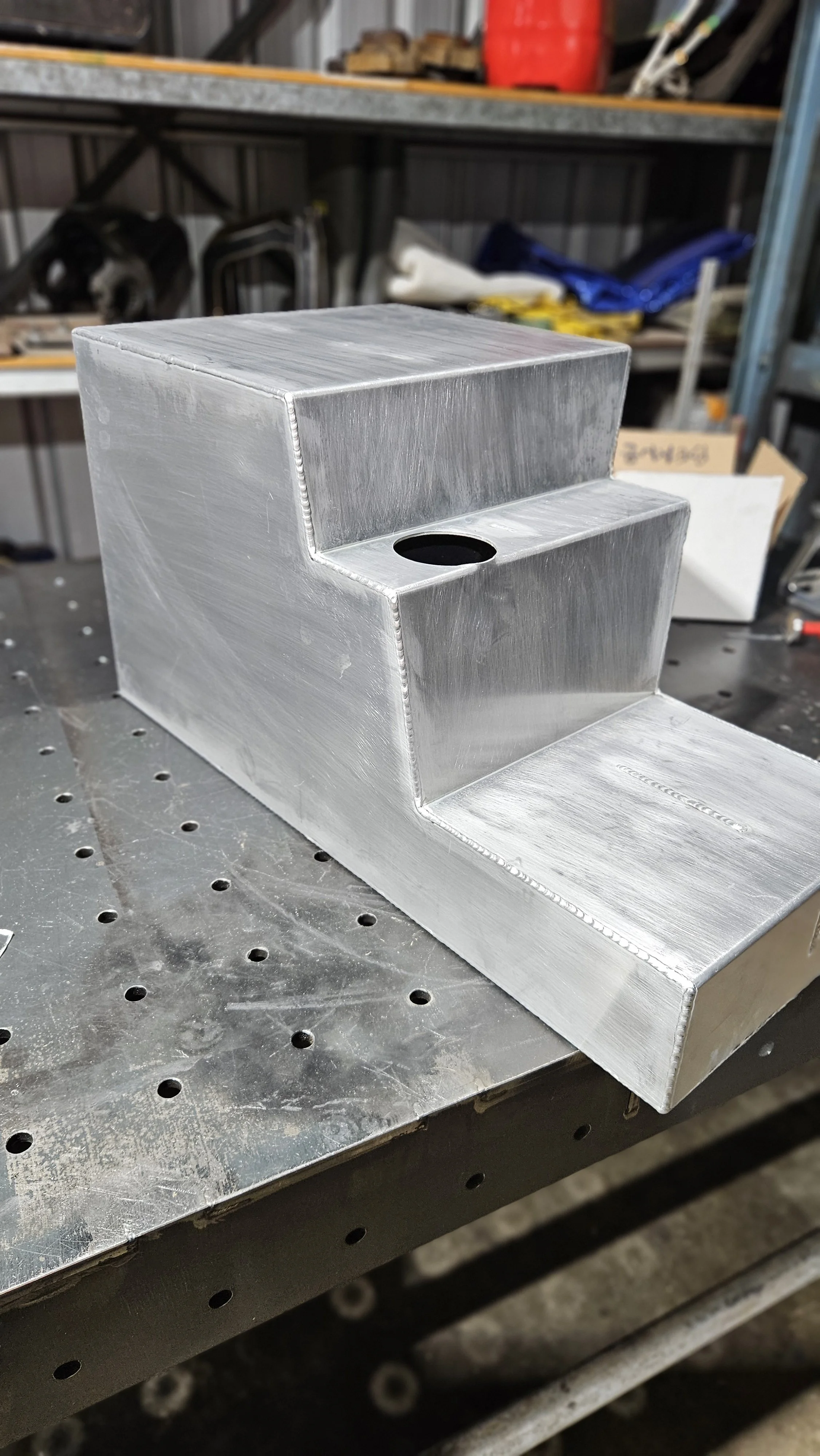 A metallic, machined metal part with a stepped and welded design, resting on a perforated work surface.