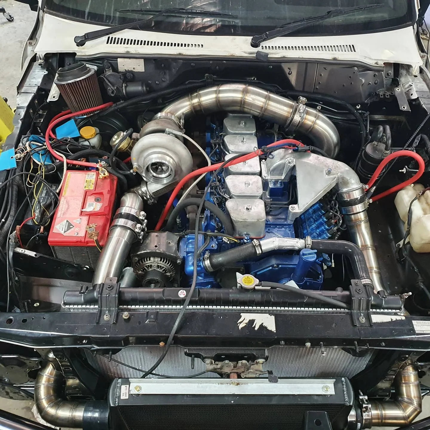 View of a car engine bay featuring a large turbocharger, red battery, various pipes, and components in vibrant colors, with wiring and parts for performance upgrade.