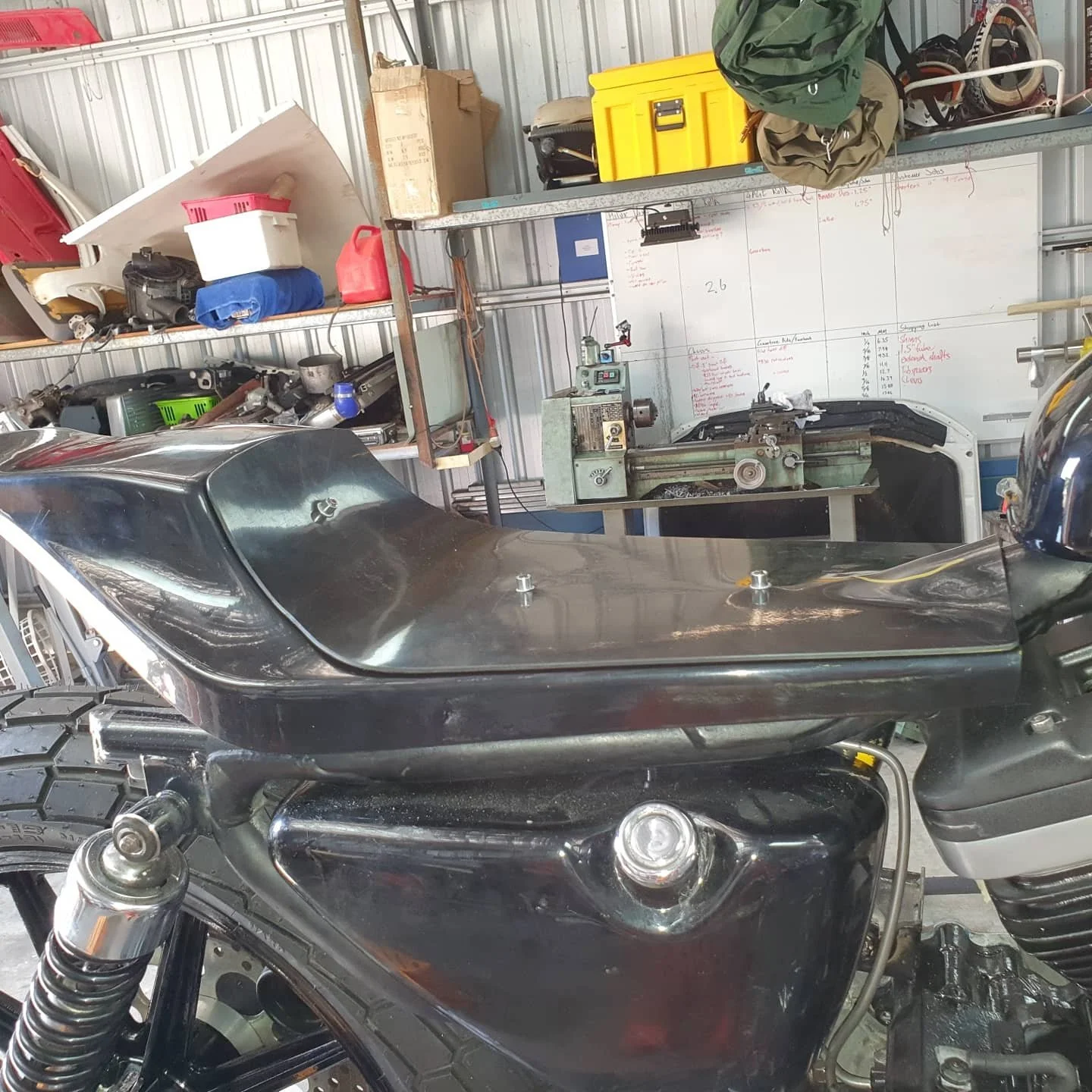 A motorcycle with a black seat in a garage, with shelves containing various tools and supplies, a lathe machine, and a whiteboard with handwritten notes in the background.