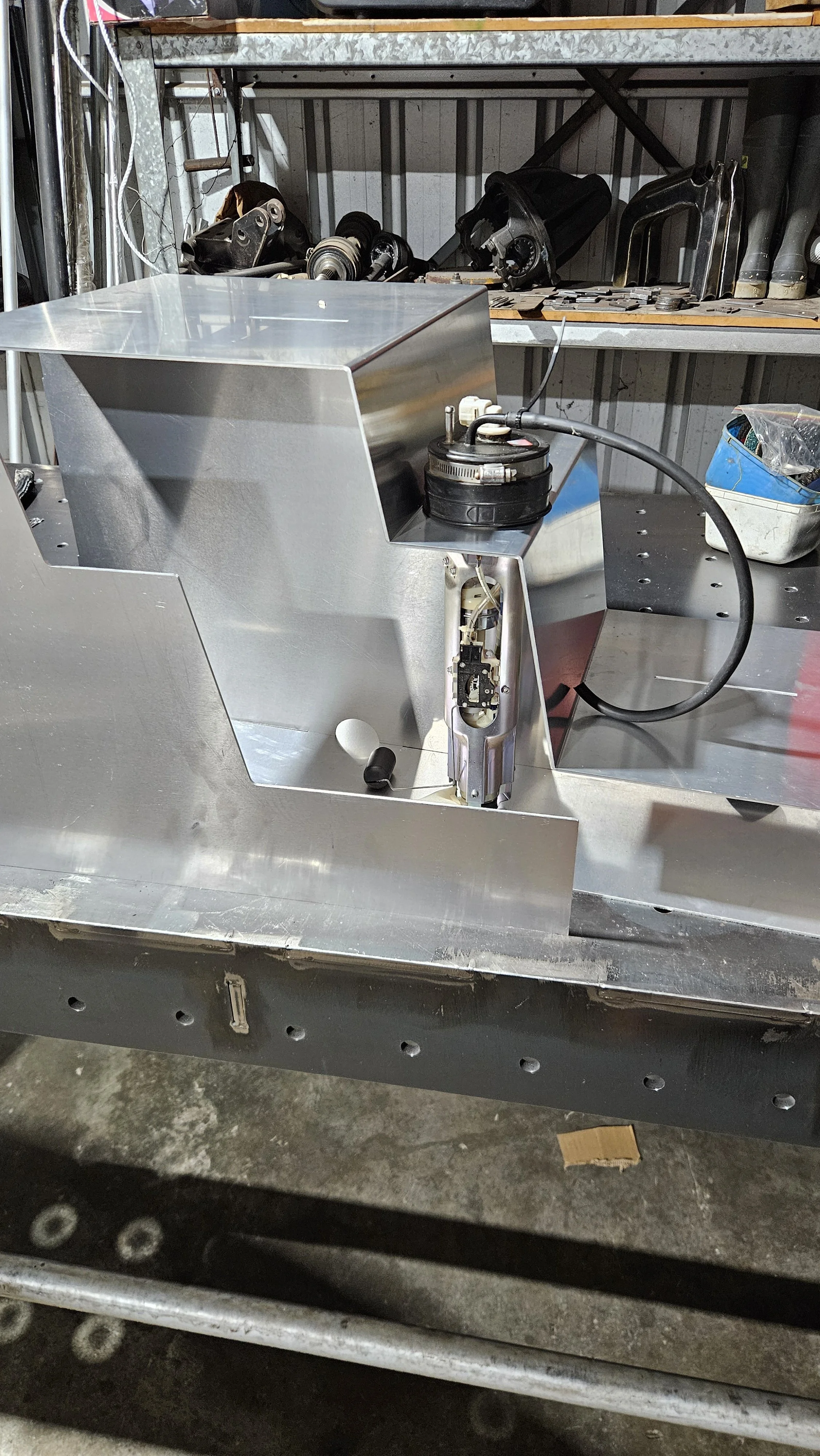 A partially assembled metal machine on a workbench in a workshop, with various tools and parts visible in the background.