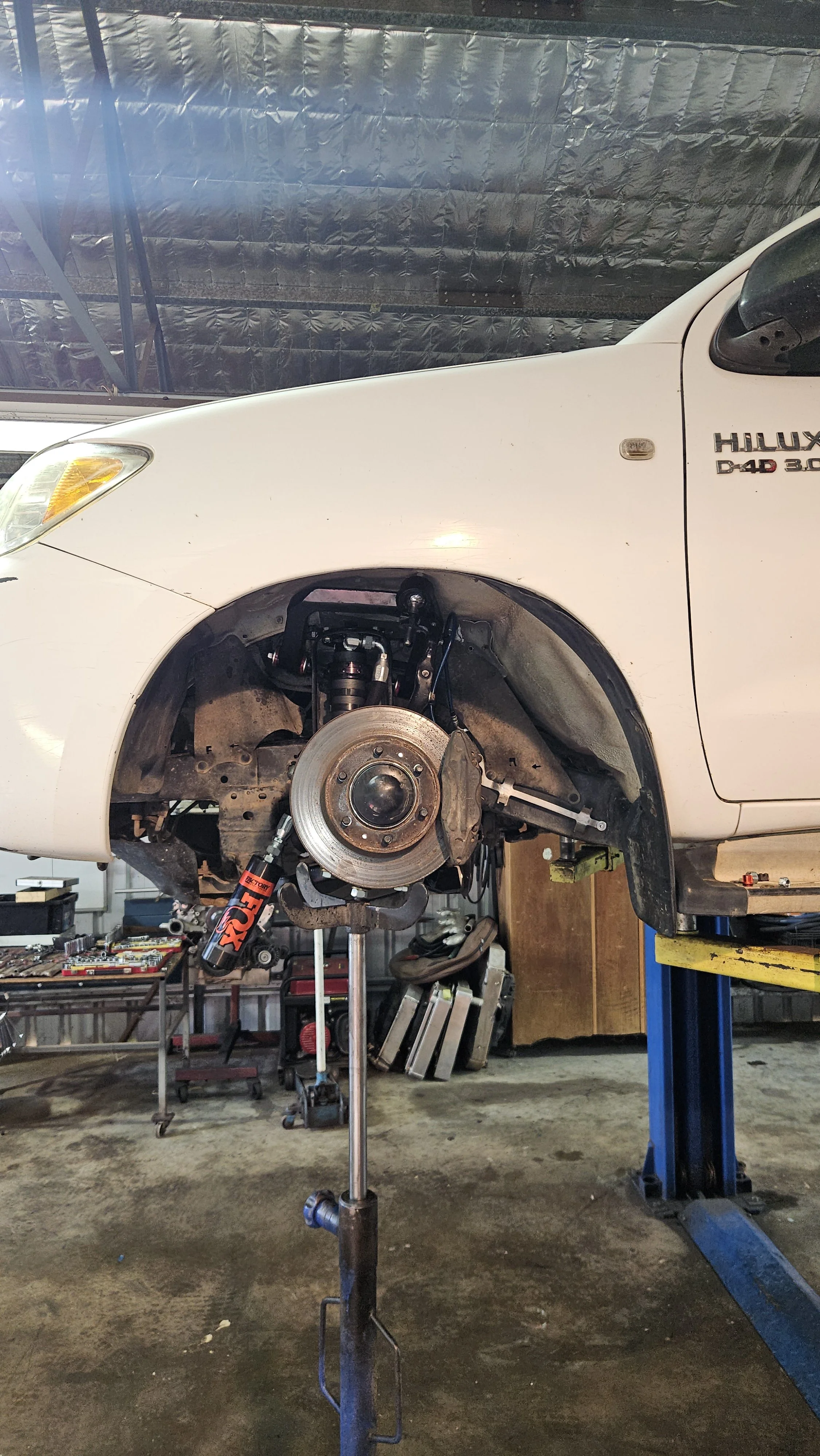 A white Toyota Hilux pickup truck raised on a lift with the front wheel removed, exposing the brake rotor and suspension components, inside an auto repair shop.
