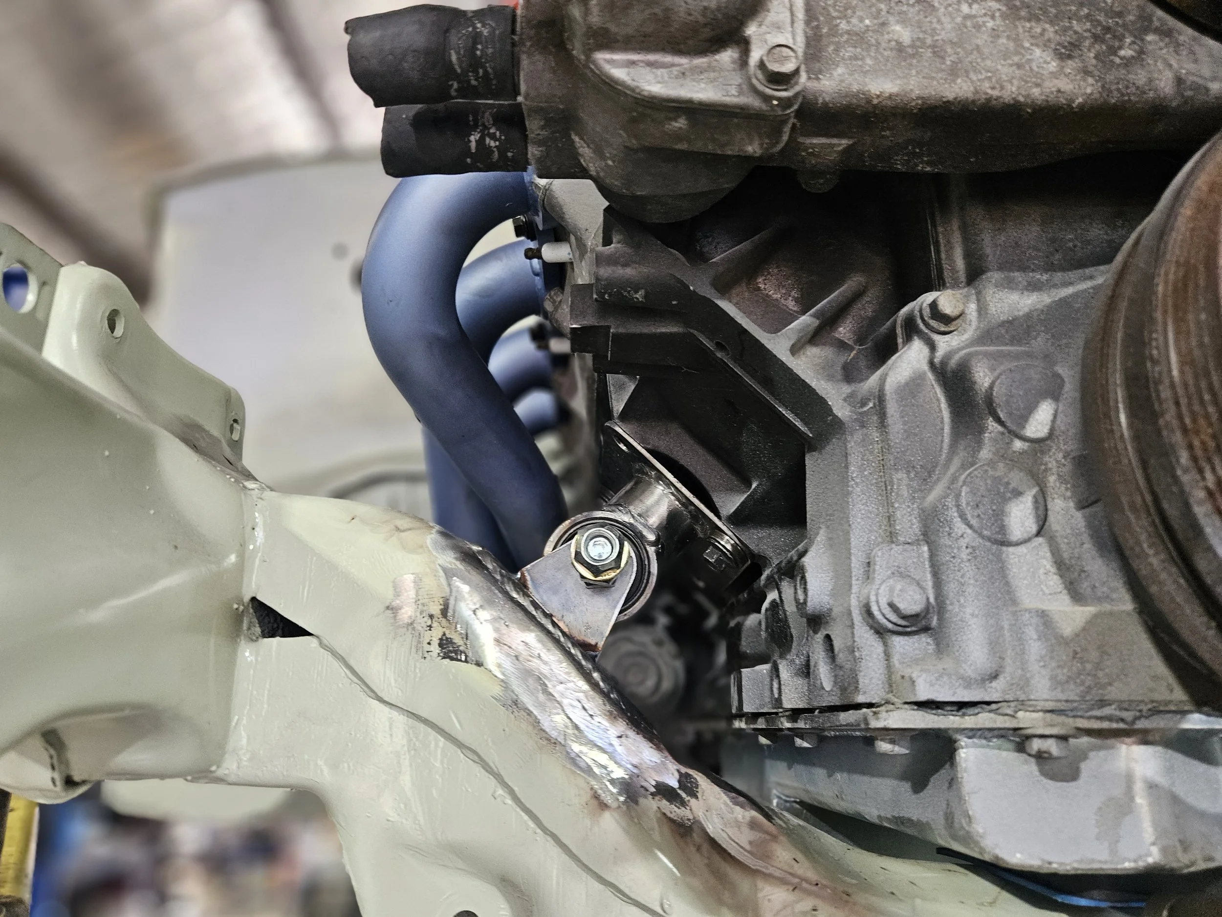 Close-up of a car engine with blue hoses connected to it, mounted on a beige metallic frame with some surface scratches, in a workshop or garage setting.