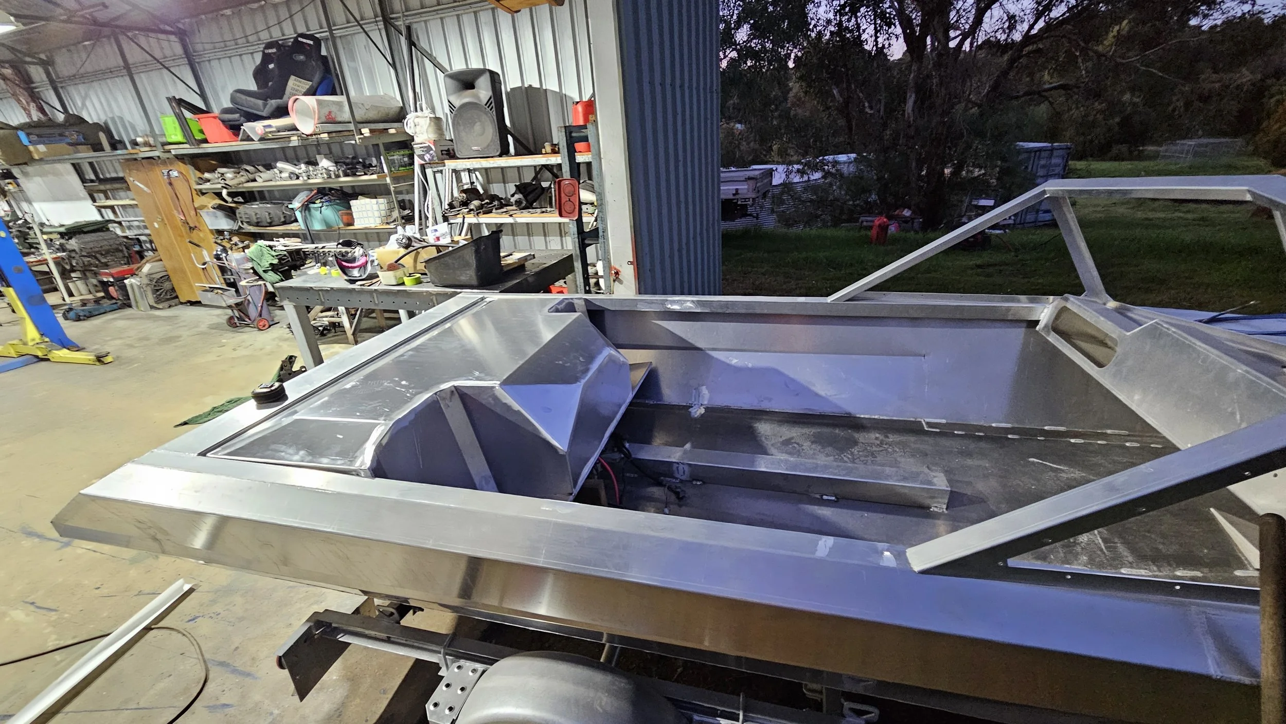Inside a garage or workshop with a boat frame in progress, surrounded by various tools and equipment on shelves and workbenches, with the outdoors visible through the open door.