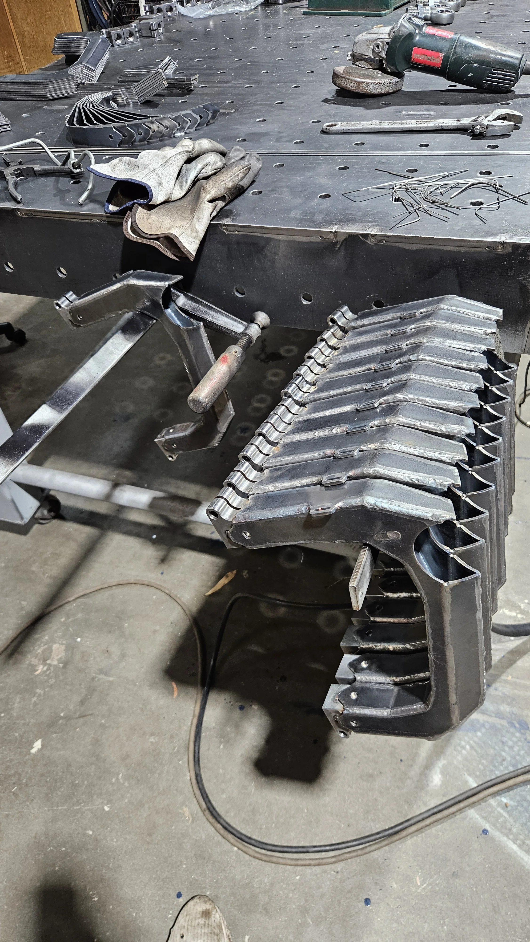 Metalwork workshop with a workbench holding various tools, gloves, and metal components in progress of fabrication or welding.