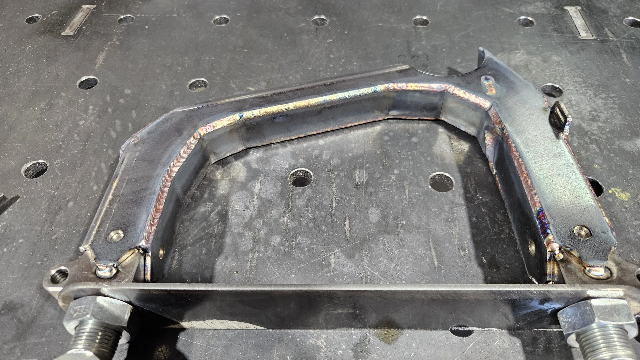 Welded metal frame on a workbench, with a rectangular shape and rounded edges, secured by bolts.