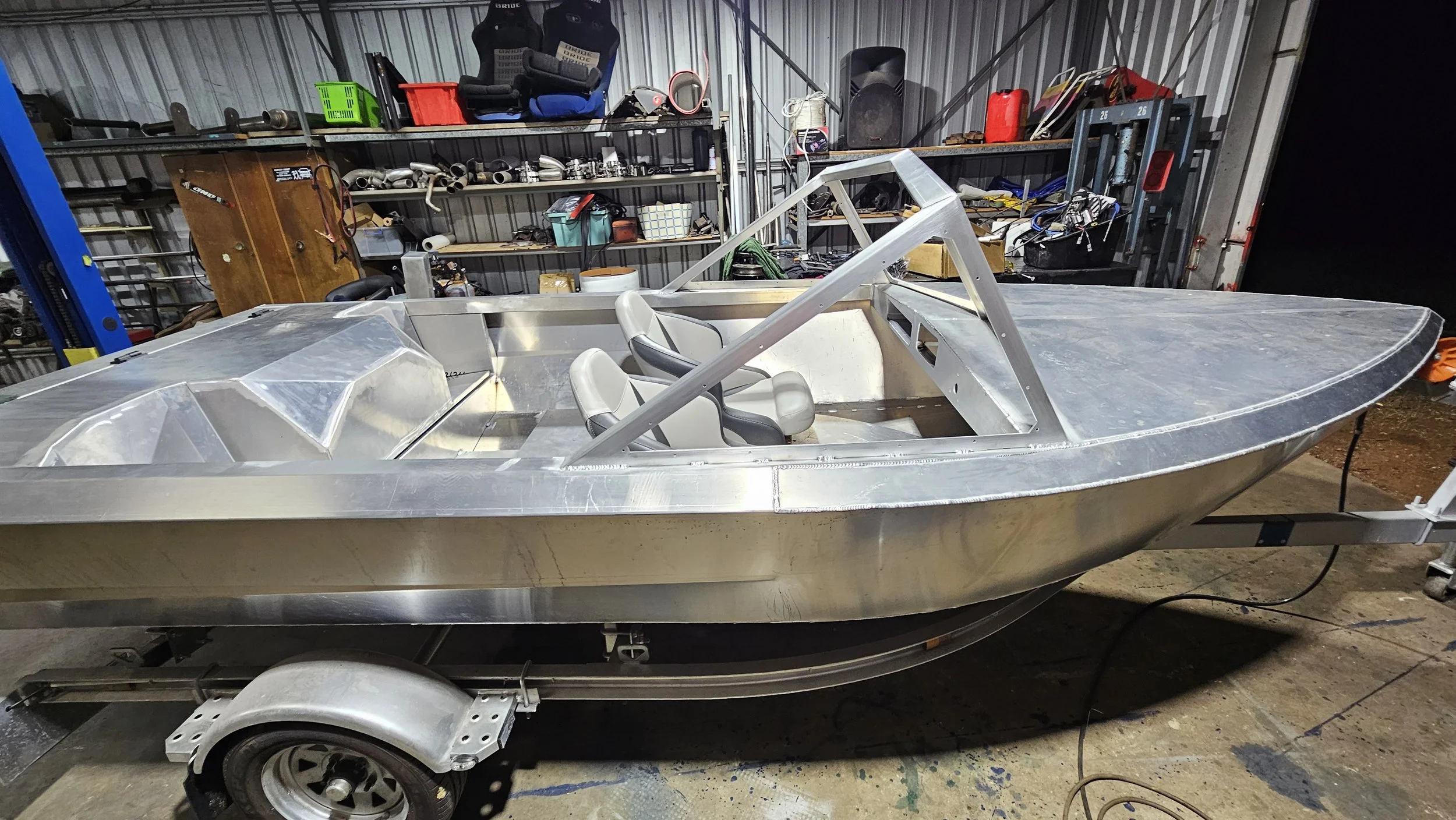 Unfinished aluminum boat with two gray and black seats inside, situated on a trailer in a workshop with shelves and tools in the background.