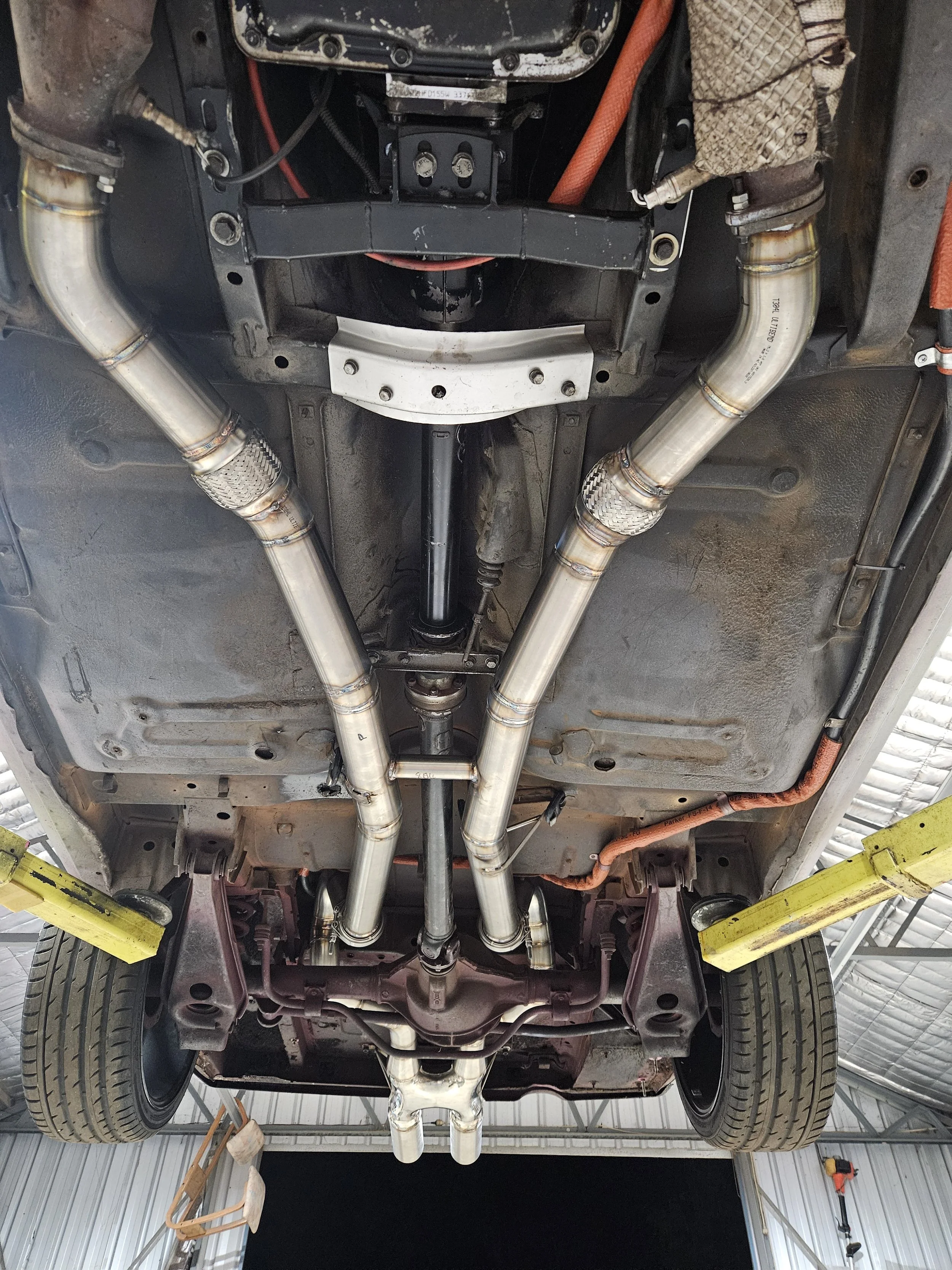 Underneath view of a car showing custom exhaust pipes, suspension, and drivetrain components in a garage or workshop.