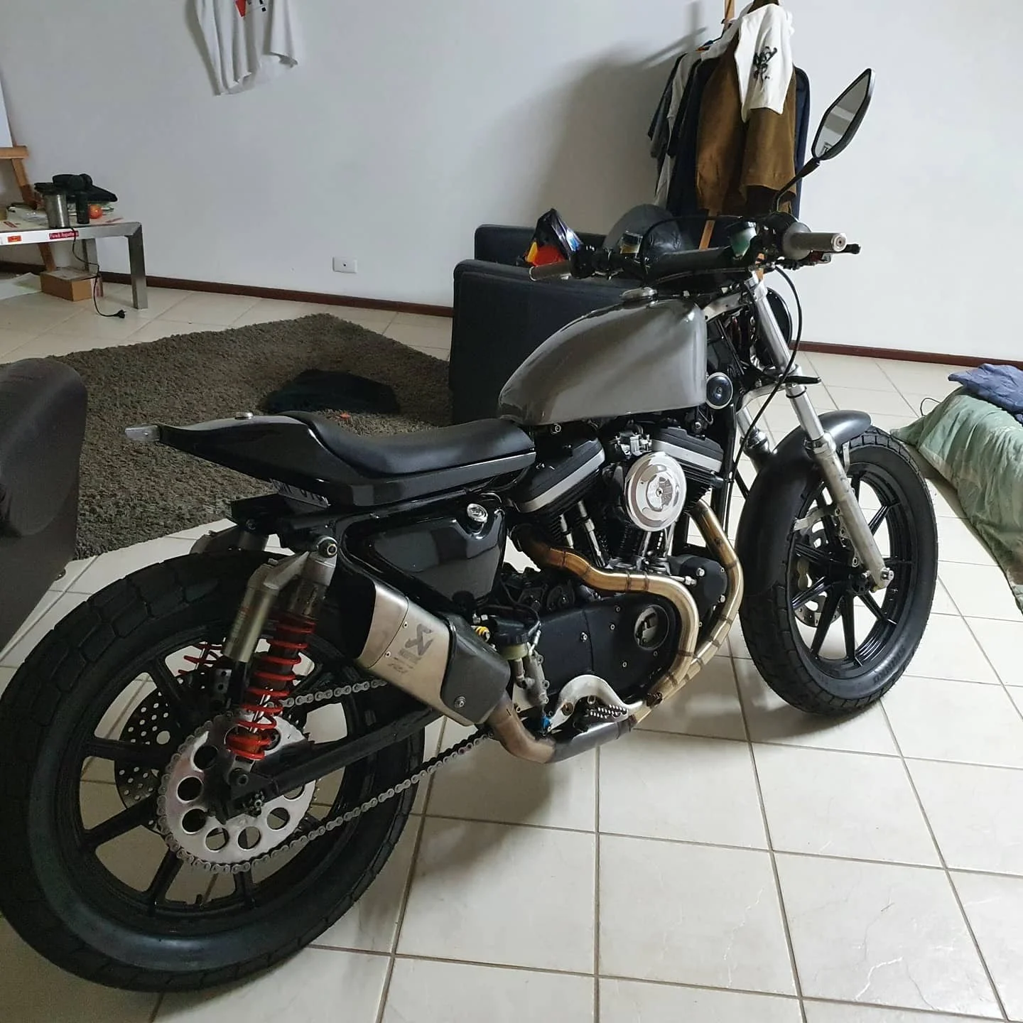 A motorcycle parked indoors on tiled floors with a living room background featuring furniture, clothing hanging, and miscellaneous items.