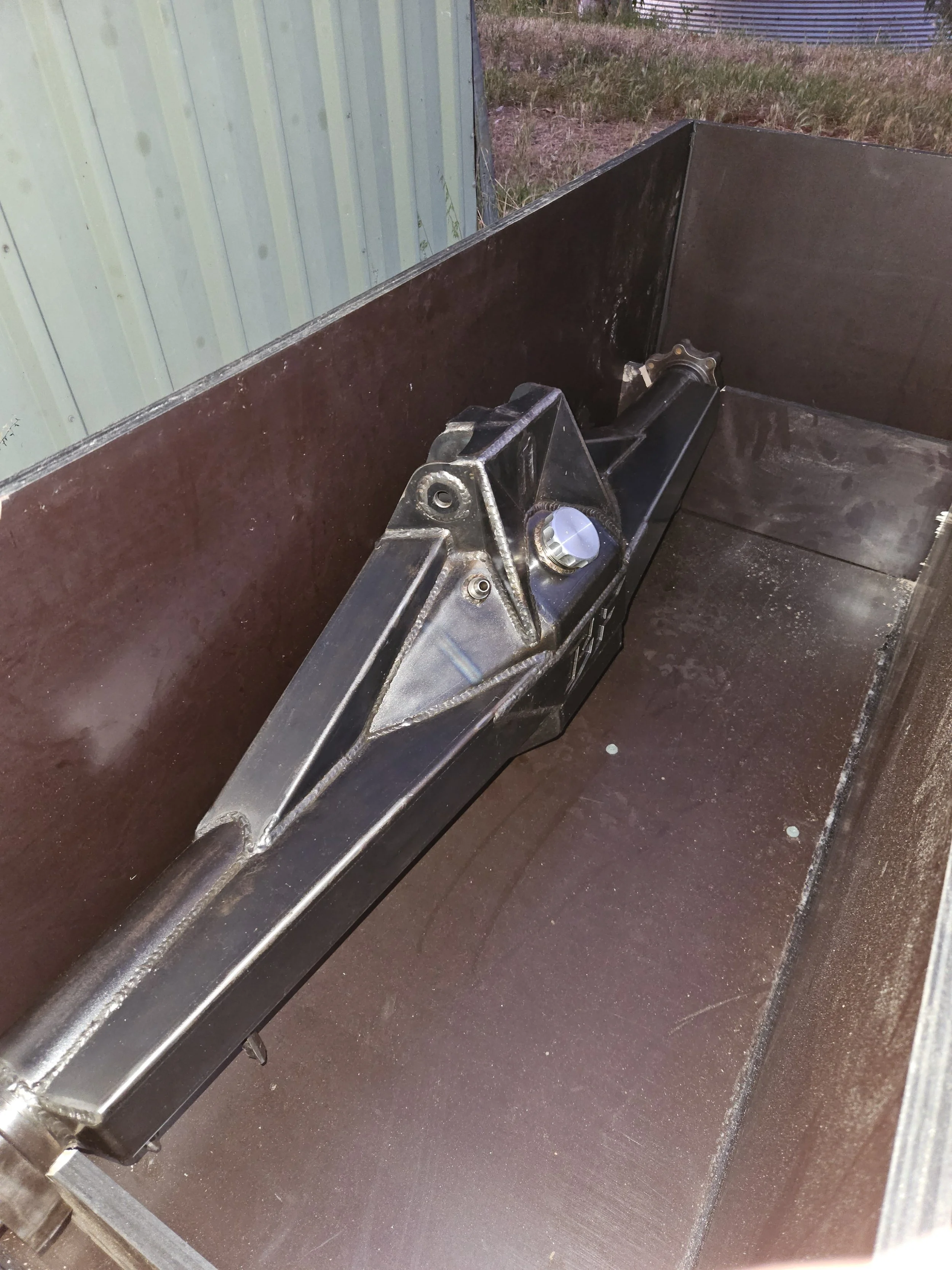 Close-up of a hydraulic tilt mechanism inside a metal container, with a metal wall on one side and a grassy area behind.