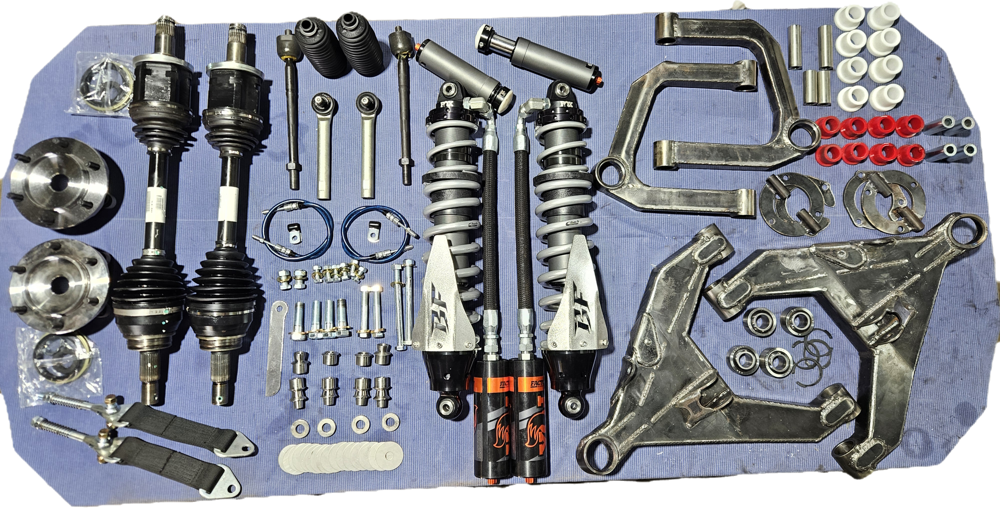 Automotive suspension parts including shock absorbers, control arms, bushings, bearings, and hardware laid out on a blue surface.