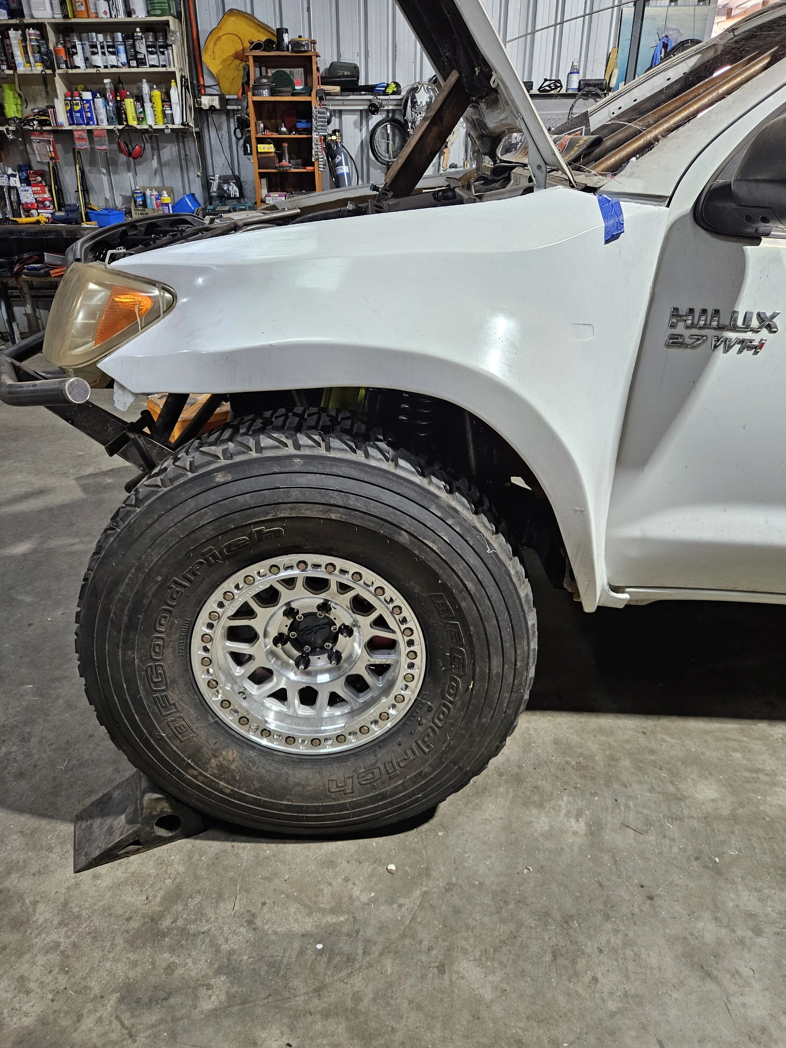 Partial view of a white Toyota Hilux pickup truck in a garage workshop, showing the front left wheel, headlight, and part of the hood with some tools and shelves in the background.