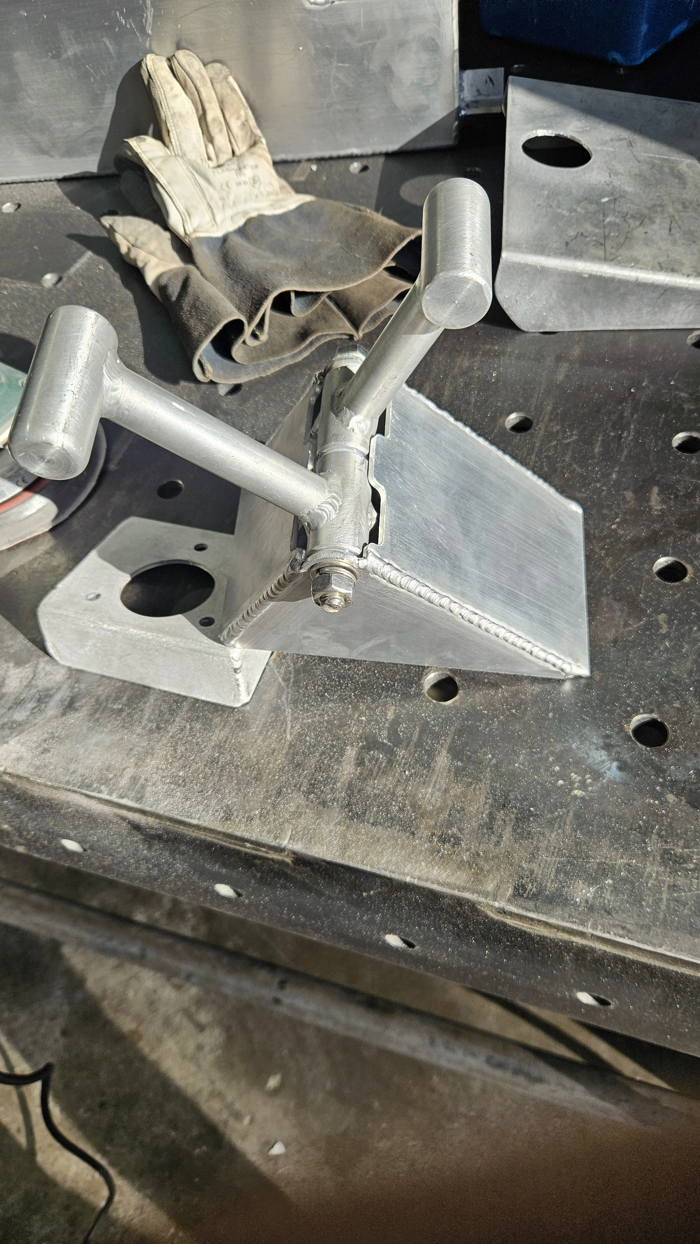 A metal workholding device with two adjustable arms on a workbench, accompanied by a pair of gloves and metal blocks.