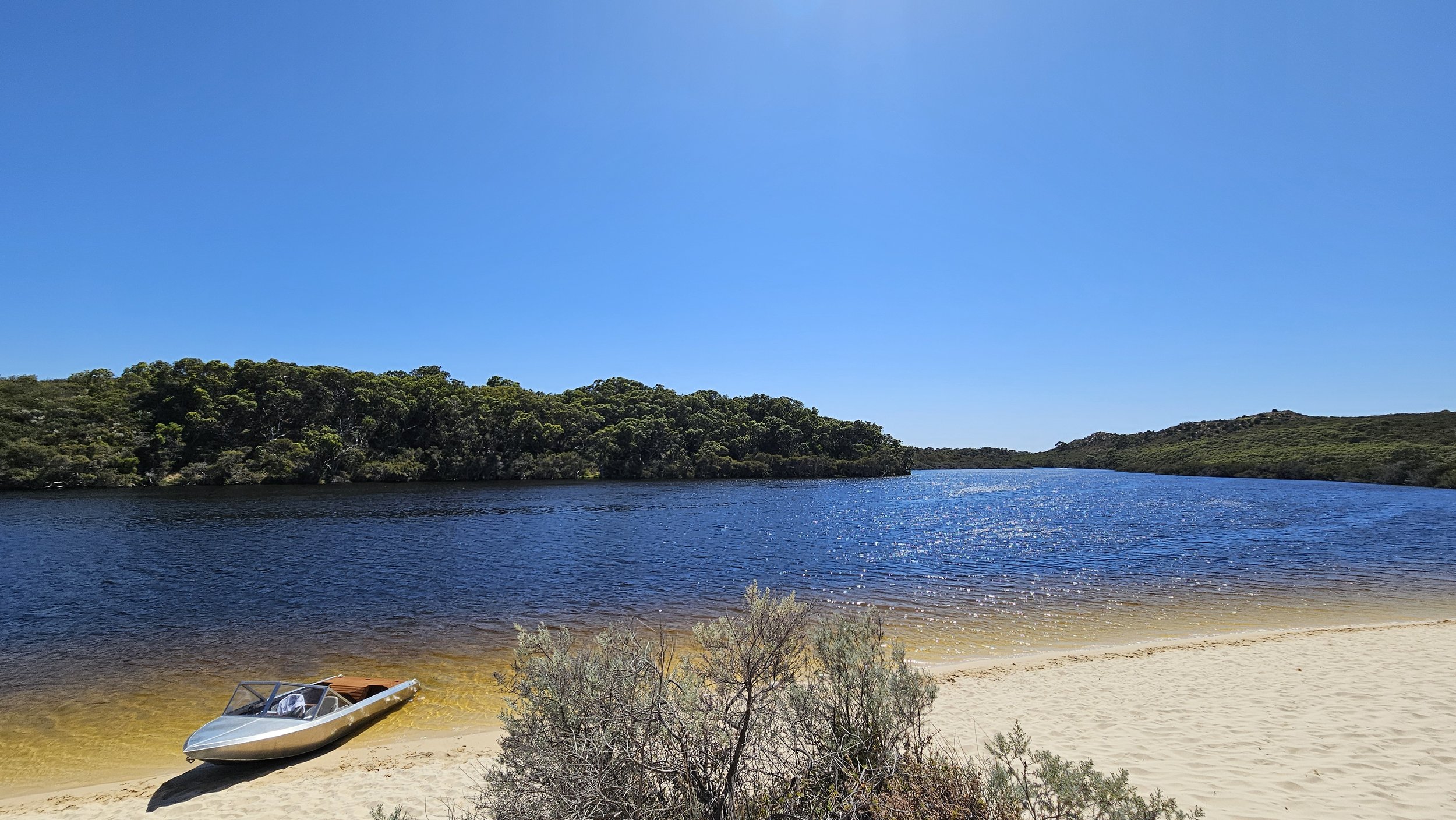 A boat resting on sandy beach by calm blue river with green trees and hills in the background under clear blue sky.