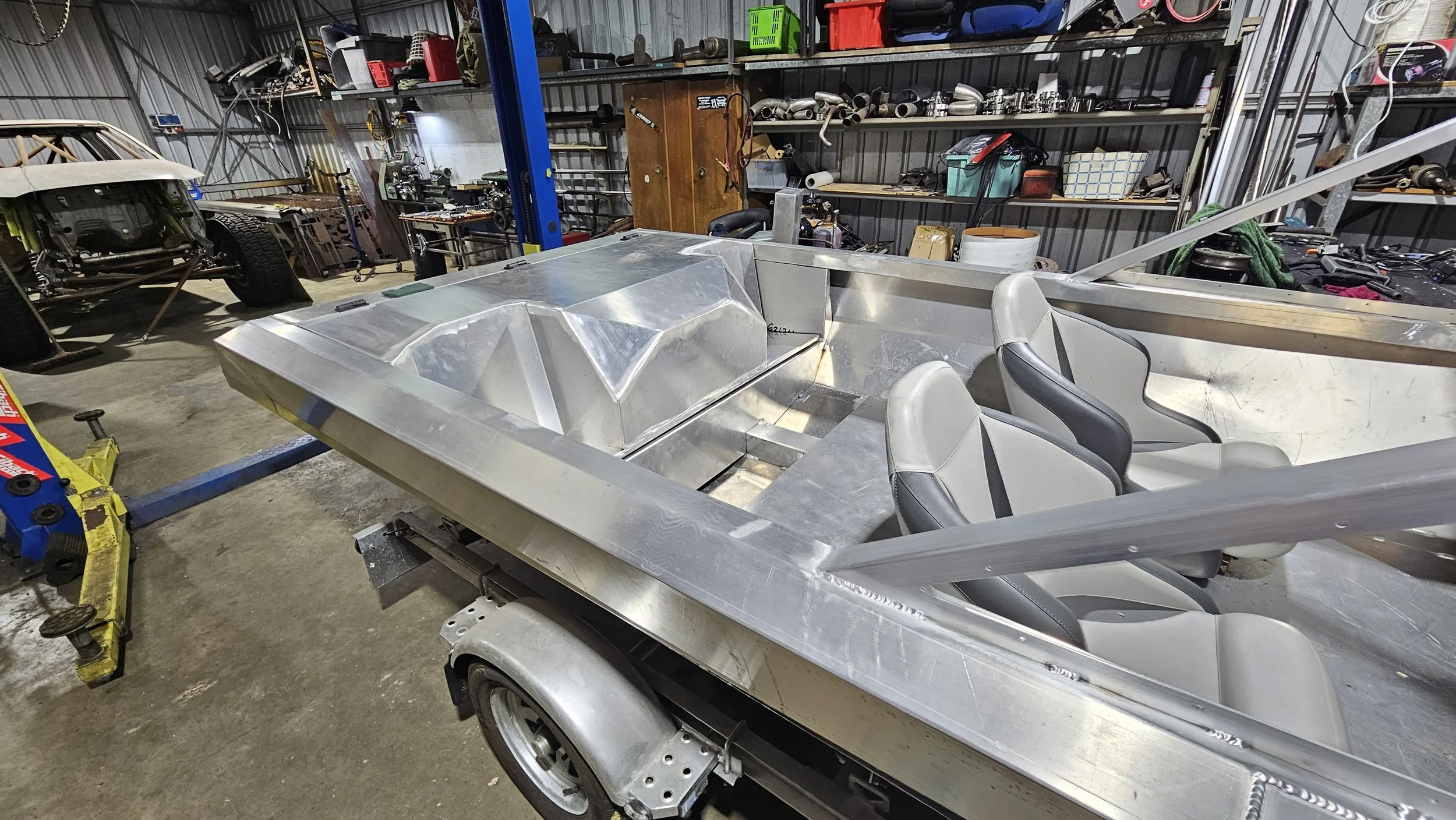 An unfinished aluminum boat with gray and black seats in a workshop. The boat is on a trailer, with tools and parts scattered around. In the background, there are car parts and a metal shelving unit with various supplies.