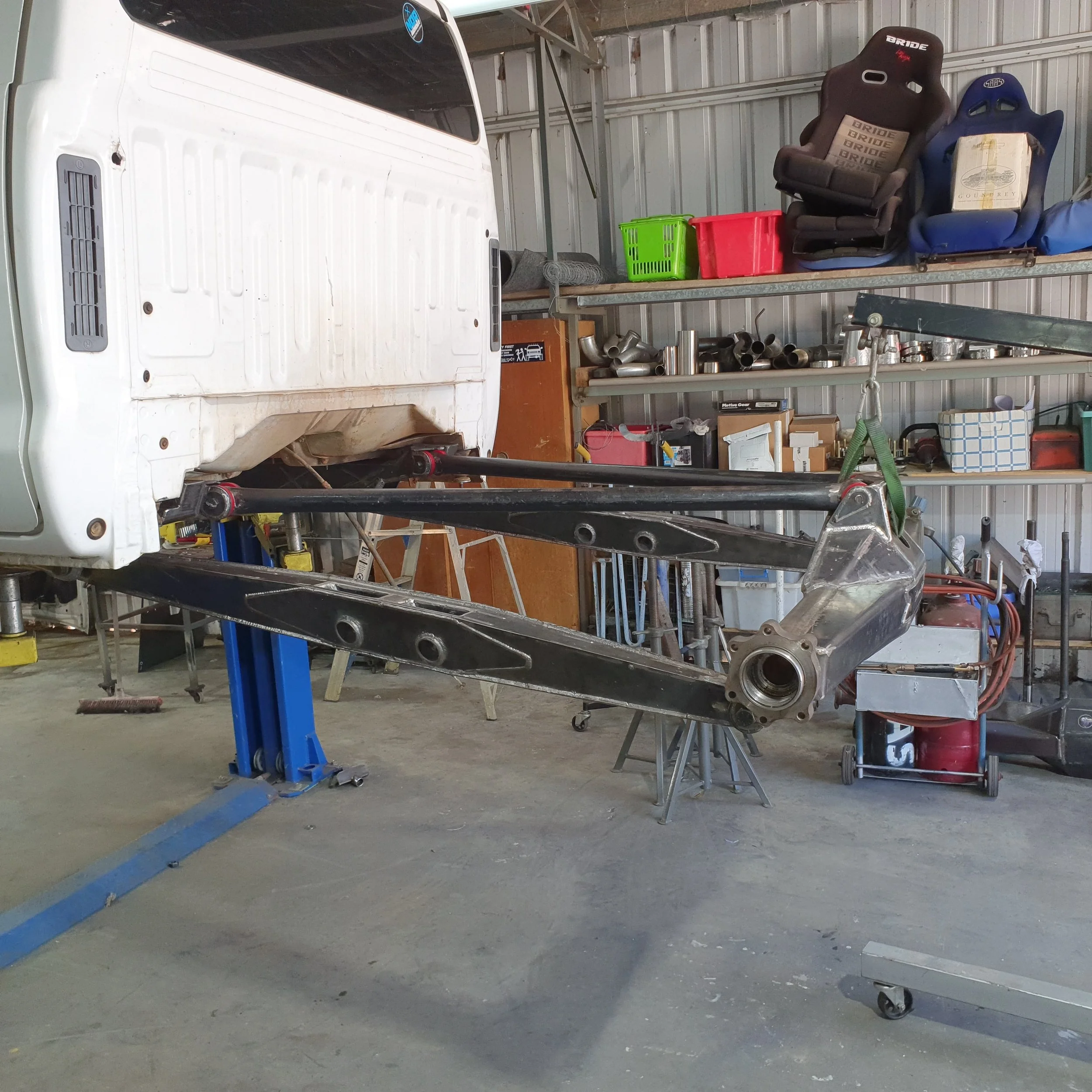 Car chassis mounted on a blue lift inside a garage with tools and car seats on shelves in the background.