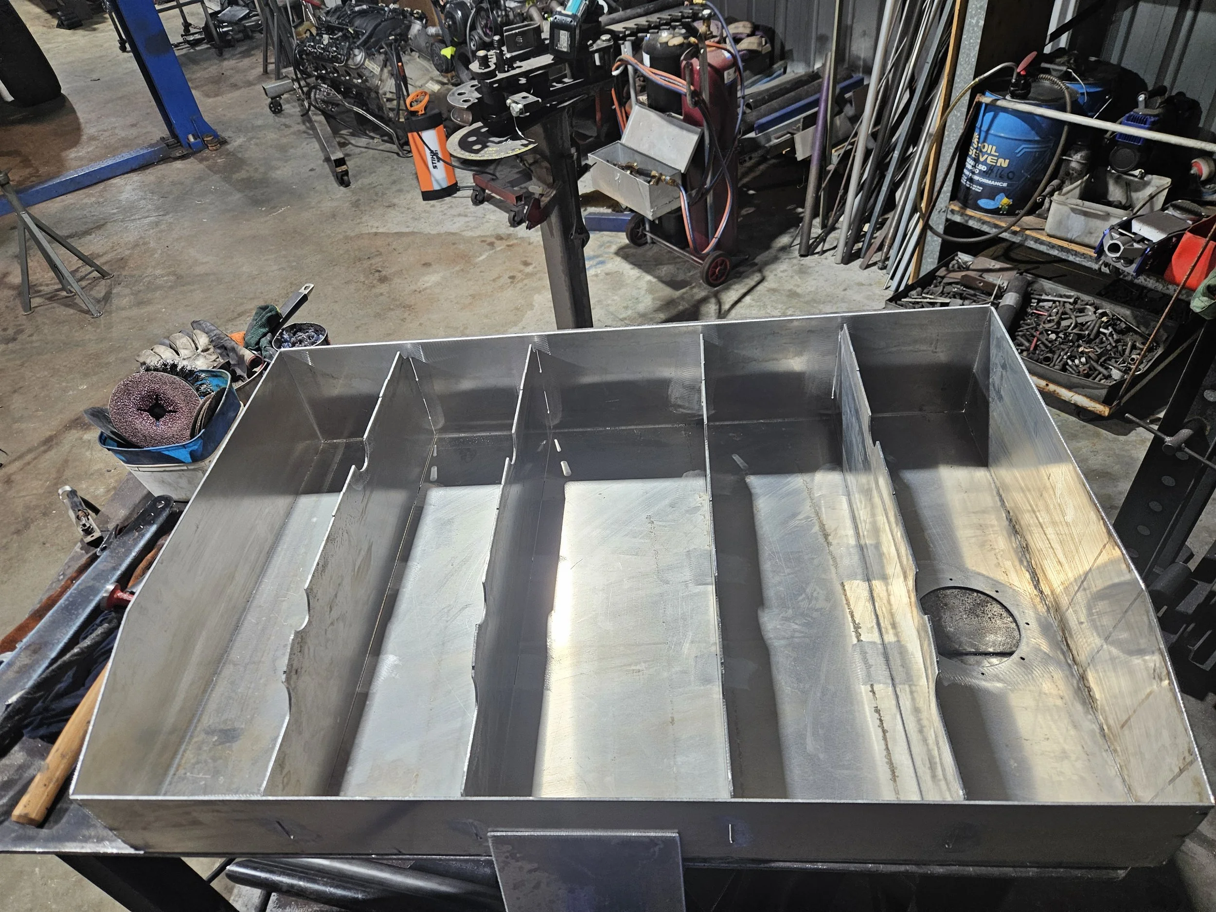 A metal fabrication workshop with a large open-top metal box on a workbench, surrounded by tools, equipment, and assorted metallic parts.