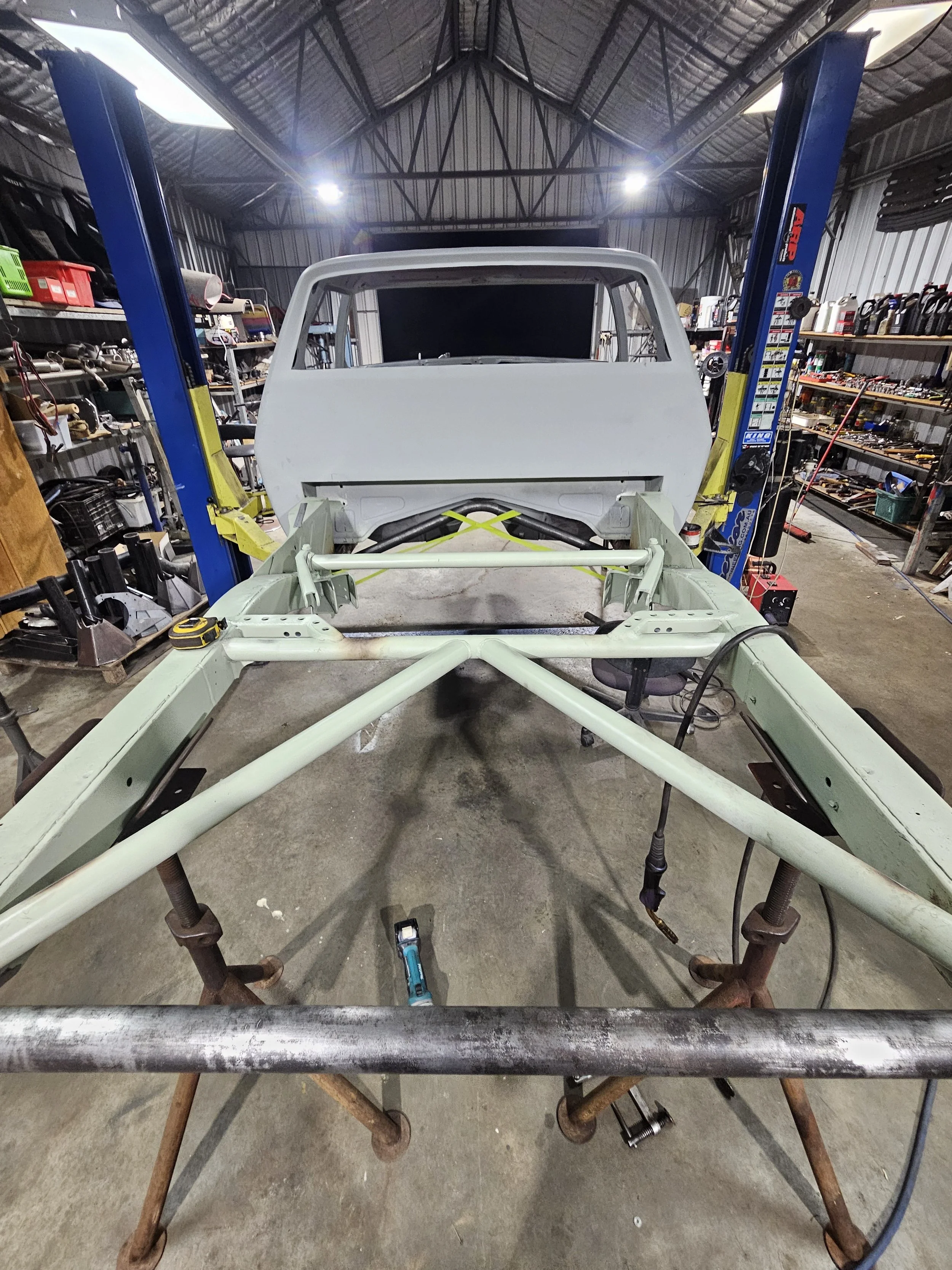 Inside a garage workshop, a vehicle frame is being assembled on a workbench, surrounded by tools and shelves with various equipment.