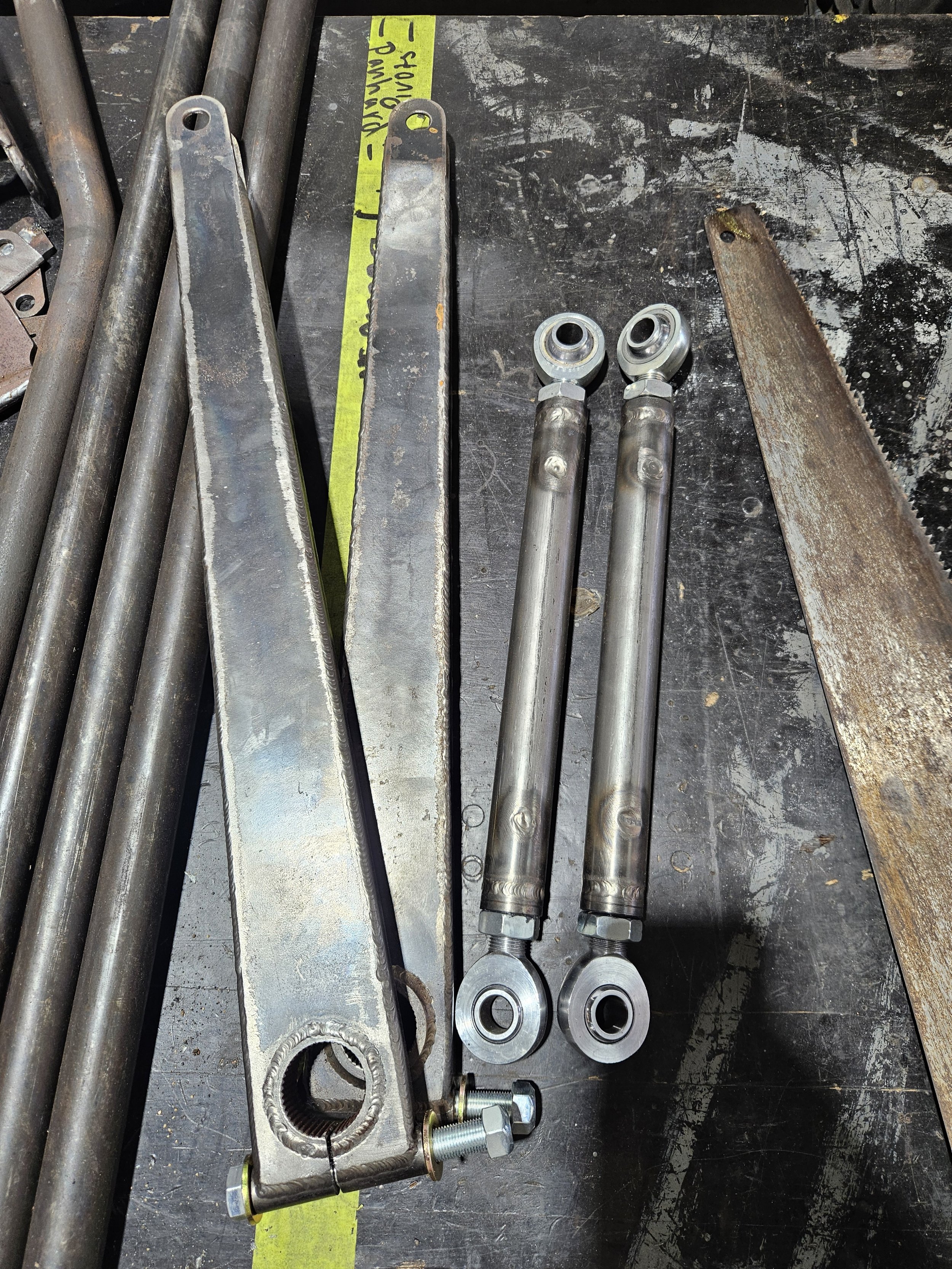 Metal workshop table with two long, cylindrical metal rods with eye-shaped ends, a large metal arm with a bolt, and a rusty saw blade.